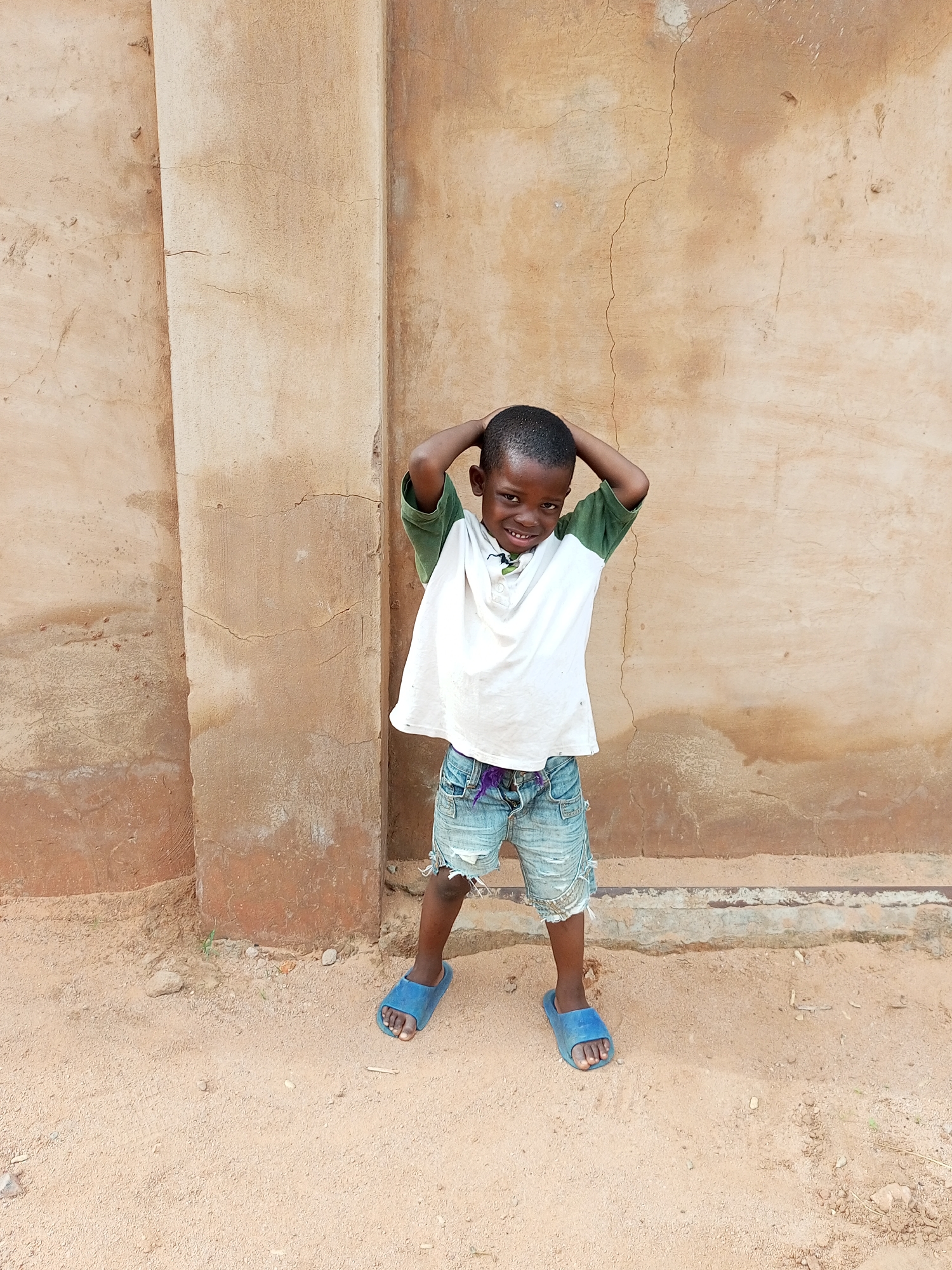A young boy stands with his hands behind his head, smiling at the camera. He is wearing a white t-shirt with green sleeves, distressed denim shorts, and blue sandals. He is positioned in front of a textured, light brown wall. The ground beneath him is sandy and appears to be outdoors. The setting is likely a part of the city of Gombe, Nigeria, given the architectural style of the wall and the sandy ground, common in many parts of northern Nigeria.
