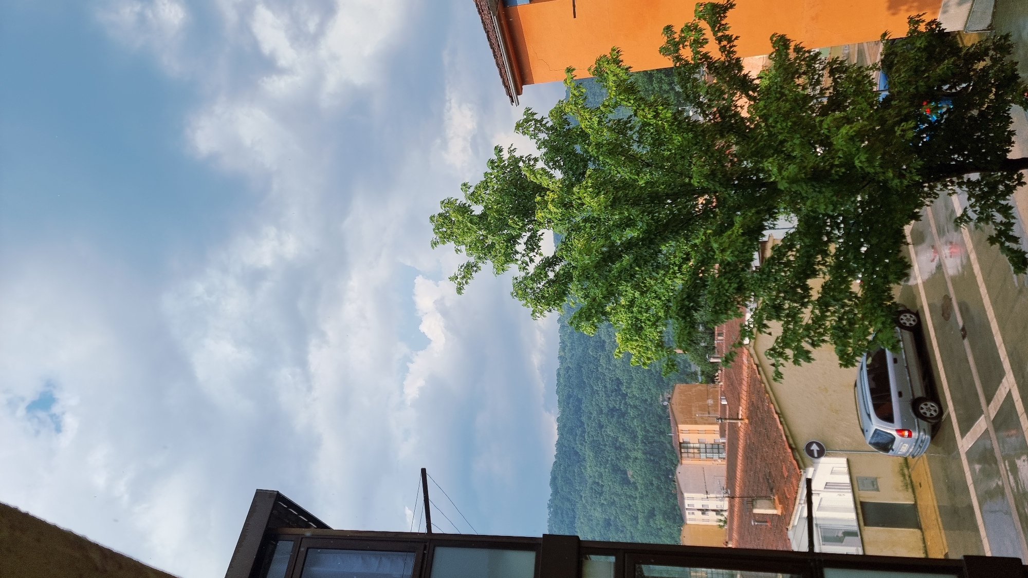 This image captures a view from a building in Sant Joan Les Fonts, Spain, looking out onto a wet street and residential buildings under a cloudy sky. A large, lush green tree dominates the center foreground, its branches full of leaves. To the left, the dark brown exterior of the building from which the photo is taken is visible, featuring a large window.
In the mid-ground, a silver minivan is parked on the wet, patterned pavement, reflecting the overcast sky. Behind the minivan, several buildings with terracotta roofs are visible, including a cream-colored building and other structures with varied architectural styles and colors. A blue traffic sign with a white arrow indicating direction is mounted on a pole near the minivan.
Further in the background, a densely forested hillside rises, covered in green foliage. The sky is filled with a mix of gray and white clouds, with a small patch of blue visible towards the top left. The overall impression is a somewhat damp and overcast day in a European town.