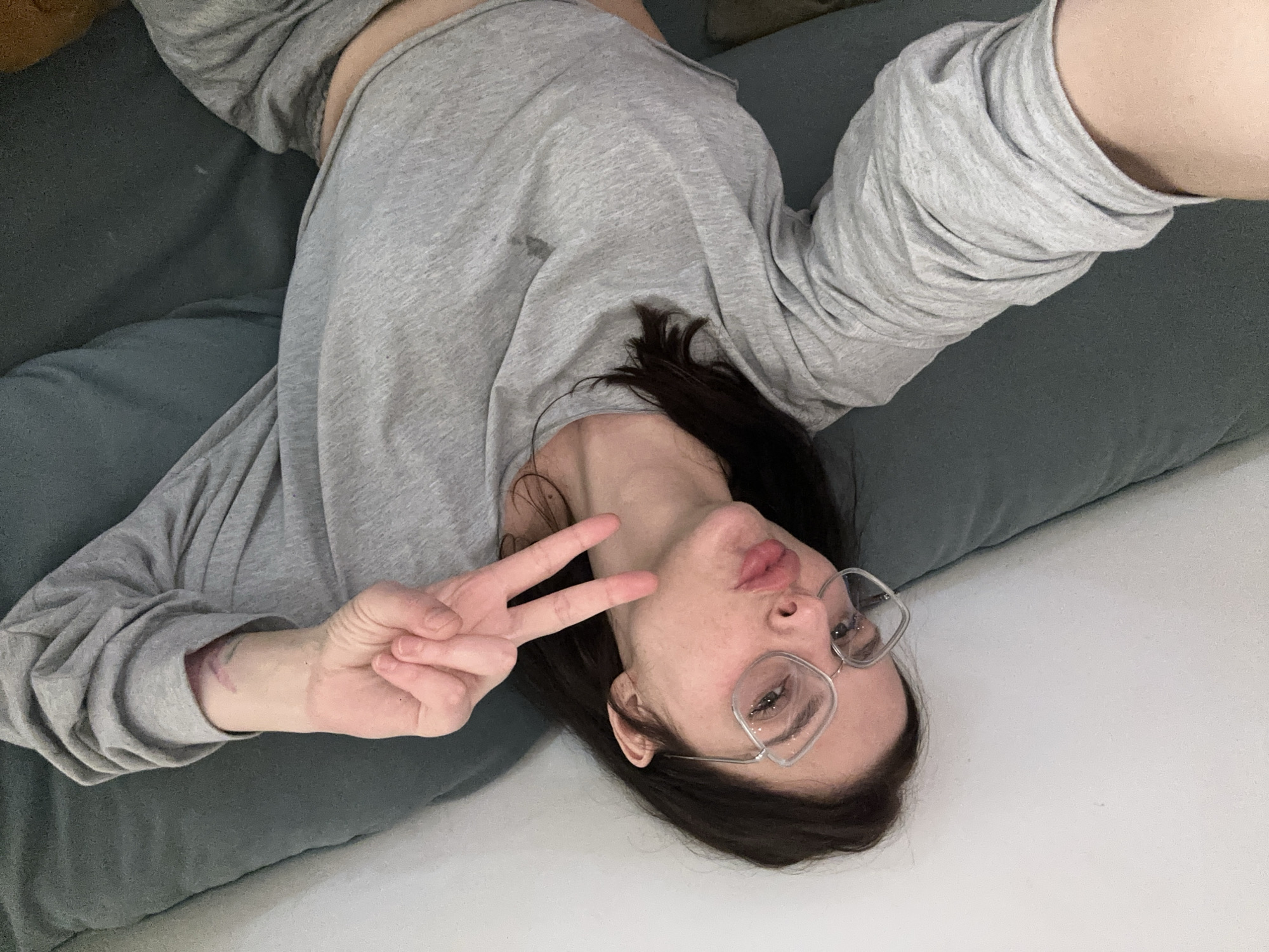 The image shows a person lying on a couch, taking a selfie. The person is wearing a grey long-sleeved shirt and glasses. They are making a peace sign with their left hand and puckering their lips. The background is a couch and a white wall. The lighting appears to be artificial, suggesting it could be indoors at any time of day. The person's expression is playful and relaxed. There is no visible text in the image.