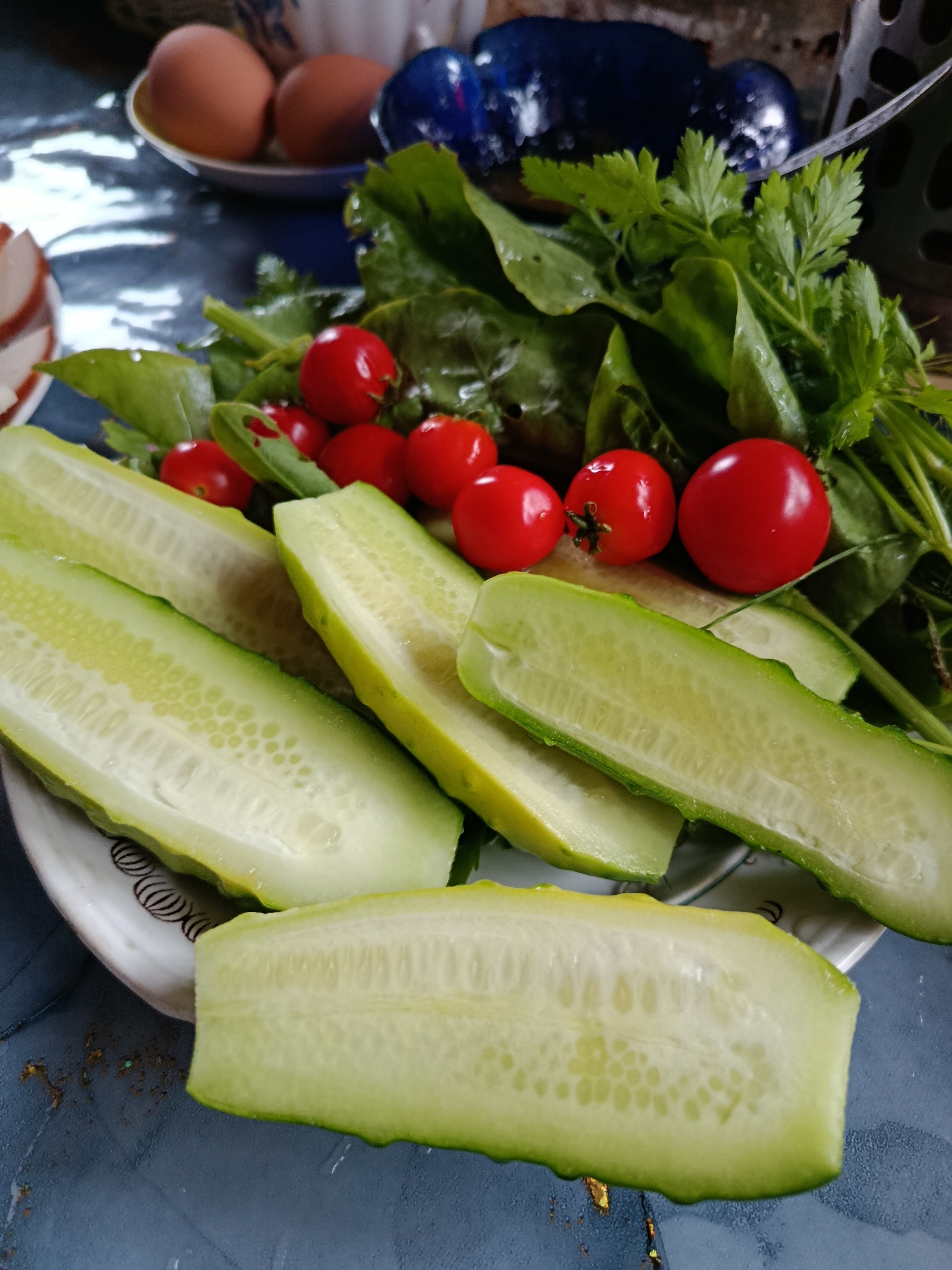 In an indoor setting, possibly a kitchen or dining area in Verezya, Russia, a close-up shot focuses on a platter of freshly prepared ingredients for a meal. The platter holds several elongated, sliced cucumbers, their pale green flesh revealing a lattice of small seeds. Scattered amongst them are bright red cherry tomatoes and a generous bunch of leafy green vegetables, including spinach and parsley. The ingredients are arranged on a white plate with a decorative rim, resting on a dark, textured surface. In the background, out of focus, are two brown eggs in a small dish and glimpses of dark blue ceramic objects, hinting at other items on the table. The lighting suggests a natural light source, casting subtle shadows and highlighting the vibrant colors of the produce. The overall scene conveys a sense of a simple, wholesome meal being prepared.