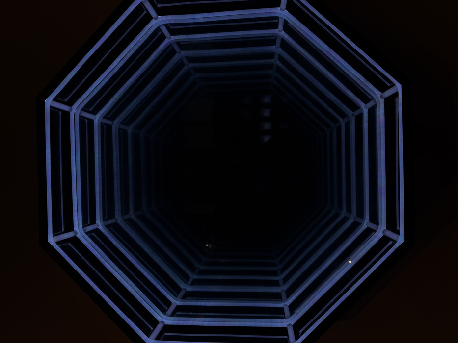 The image displays an octagonal infinity mirror installation, located within the Sense Hotel Sofia, Bulgaria. The central subject is a repeating series of blue-purple illuminated octagonal frames that recede into a deep, dark central void, creating an optical illusion of infinite depth. The outermost frame is clearly defined, with subsequent frames diminishing in size. The primary illumination originates from these glowing frames, casting a blue-purple hue against the predominant blackness of the perceived tunnel. A single, distinct, bright yellowish-white point of light is visible within the lower-left section of the innermost clear octagonal frame. Further into the dark void on the right, several faint, indistinct dark rectangular elements are subtly visible. The scene is static, devoid of people, subjects, or direct actions.