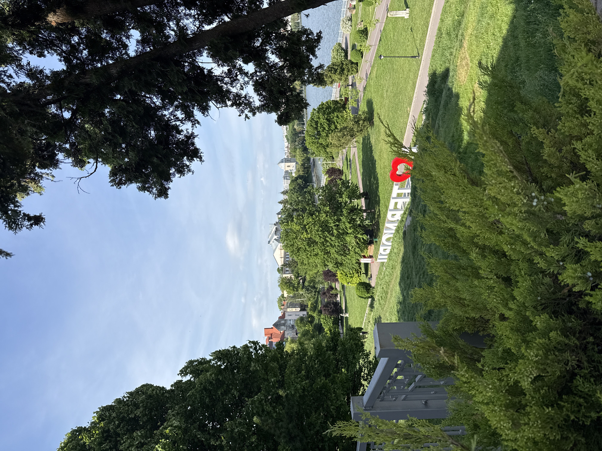 The image shows a park in Ternopil, Ukraine. In the foreground, there's a view through trees and a railing, leading to a grassy park area. A large white sign with a red heart is visible, likely a public art installation spelling out "I <3 Ternopil" in Ukrainian. Beyond the park, buildings and possibly a body of water are visible in the background. The sky is blue with some clouds.
