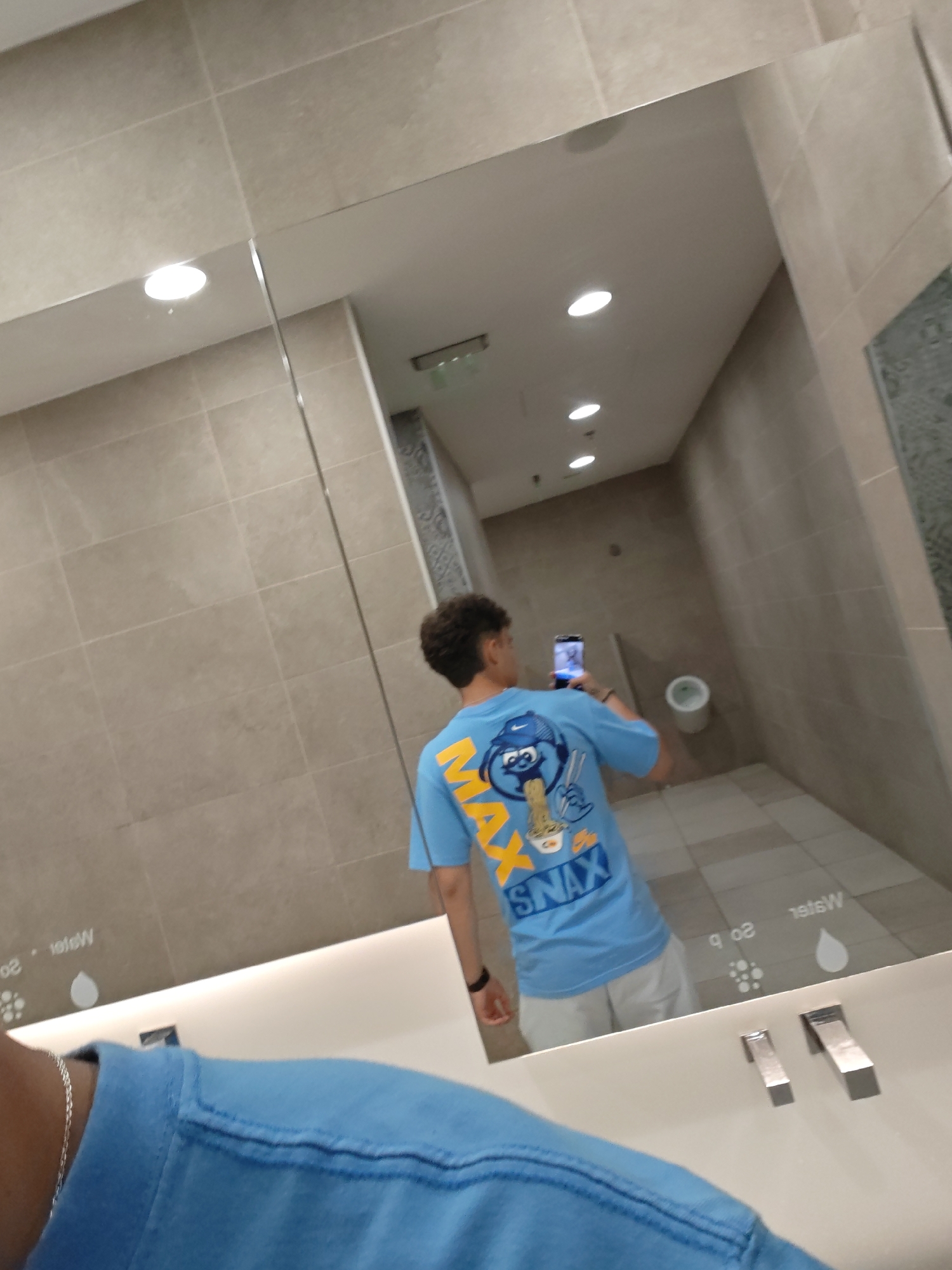 The image is a reflection in a large, commercial restroom mirror. A young person with dark, curly hair is taking a selfie with their phone. They are wearing a light blue t-shirt with a cartoon graphic of a character eating ramen, and the word "SNAX" in yellow lettering. They are also wearing light-colored shorts and a black watch.
The restroom appears to be modern, with beige tiled walls and floors. There are several recessed lights in the ceiling. In the reflection, a white toilet is visible in the background. The sink area features a white countertop with two modern, brushed metal faucets. On the mirror, near the sink, there are subtle etched markings, including a droplet symbol and the word "water". The overall environment suggests a public space such as a mall, airport, or large office building. The location is identified as Stara Zagora, Bulgaria.