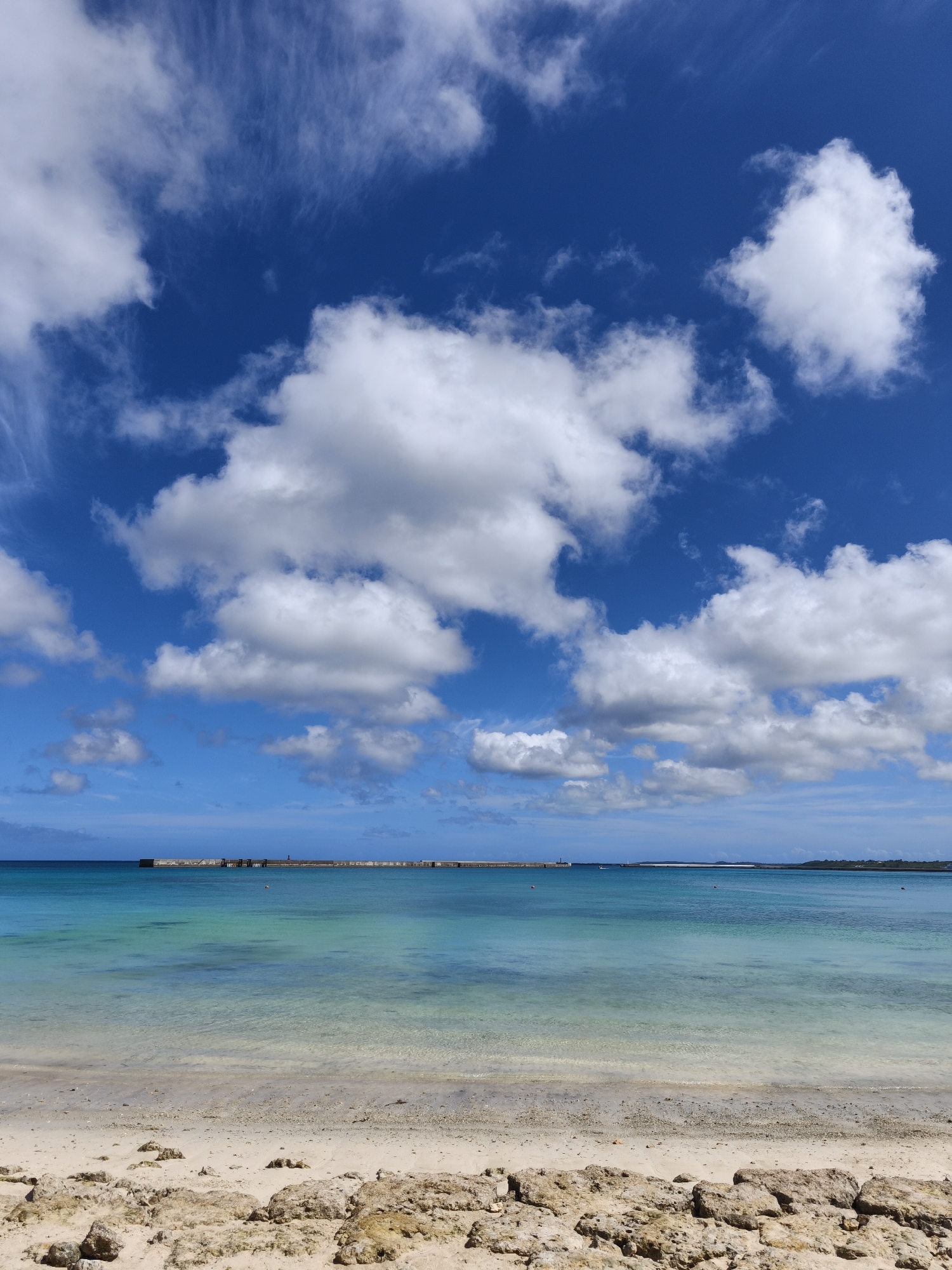 The image depicts a serene beach scene in Miyakojima, Japan, under a vibrant blue sky dotted with fluffy white clouds. The foreground features a sandy beach scattered with rough, light-brown rocks. Beyond the beach, the crystal-clear turquoise water of the ocean stretches to the horizon, gradually deepening in color. A long, man-made structure, possibly a breakwater or pier, is visible in the distance, with a lighthouse or tower at one end. Small orange buoys are also scattered across the water. The overall impression is one of tranquility and natural beauty, characteristic of Miyakojima's coastal landscapes.