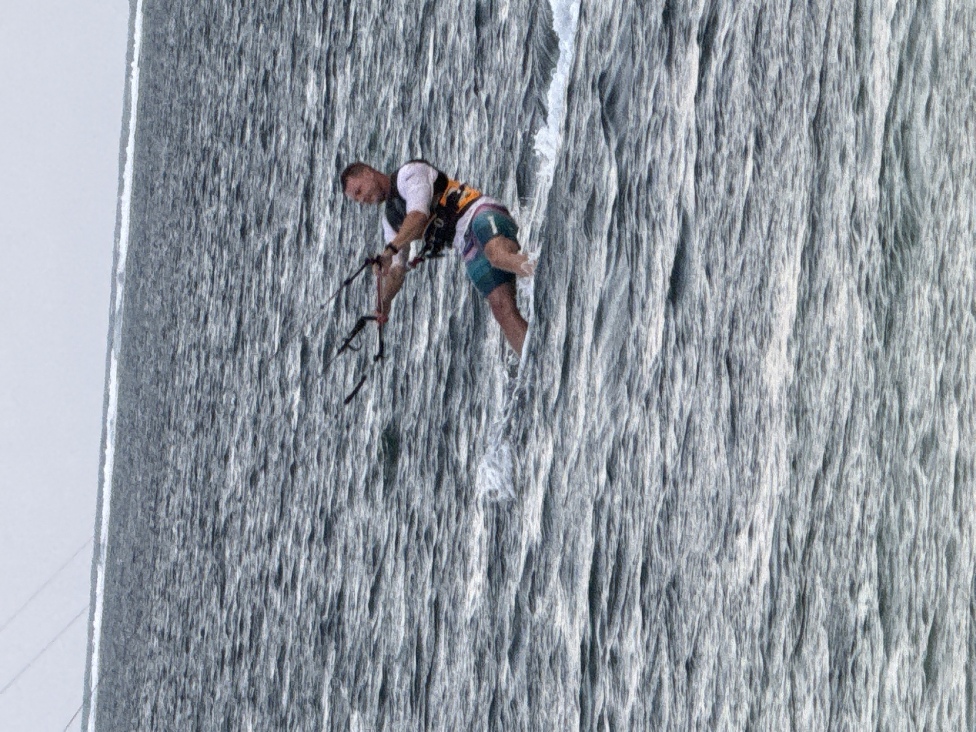 A man kitesurfs on the choppy, grey ocean. He is wearing a white t-shirt, a black and orange life vest, and teal and pink patterned shorts. His body is angled, and he holds the control bar of his kite, which is out of frame. Water sprays around his feet as he glides across the surface. The sky is overcast and a pale grey.