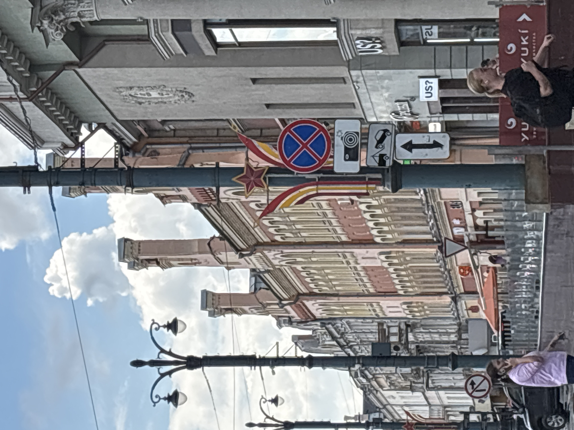 The image depicts a street scene in Novaya Usman', Russia, with an upward angle towards the sky and buildings. The sun is bright, suggesting it is daytime, and there are puffy white clouds against a blue sky.
The foreground features a dark lamppost with two lights. Behind it, ornate, multi-story buildings with detailed facades line the street. The architecture appears historical, with patterned window frames and decorative elements.
Several street signs are visible. One is a circular red and blue sign with a white cross, likely a "no stopping" or "no parking" sign. Another sign shows a camera icon, and a third displays two horizontal arrows pointing left and right within a white rectangular sign. There's also a triangular white sign with a red border.
Further down the street, a woman with blonde hair is walking away from the camera, wearing a black dress. She is holding a phone to her ear. Another person, wearing a purple top, is partially visible in the lower right corner, seemingly near a car.
On the buildings, there are signs with text. One red sign has white Cyrillic lettering that appears to say "YUUKI" with a stylized logo. Another smaller sign reads "US?".
The overall impression is of a somewhat busy urban street on a clear, sunny day.