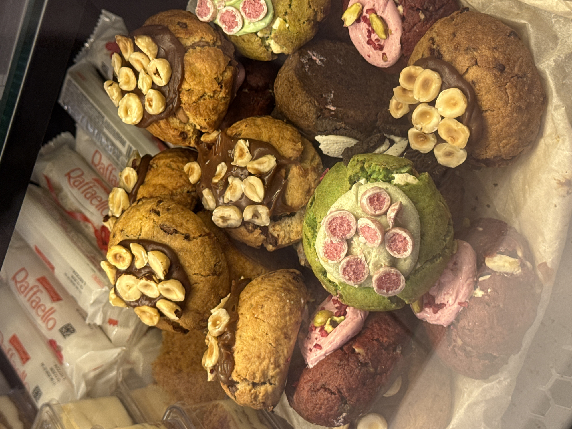 The image displays a close-up, eye-level view of an assortment of cookies and packaged candies, likely in a display case. The cookies vary in color and topping, with some featuring chocolate frosting and hazelnuts, while others are topped with pink frosting and what appear to be pieces of dried fruit or candies, and one notable cookie is green with pink candy toppings. In the background, several white rectangular packages with red lettering that reads "Raffaello" are visible. The overall lighting suggests an indoor setting, possibly a bakery or confectionery shop. The specific location within Dolgoprudny, Russia, cannot be determined from the visual information provided.