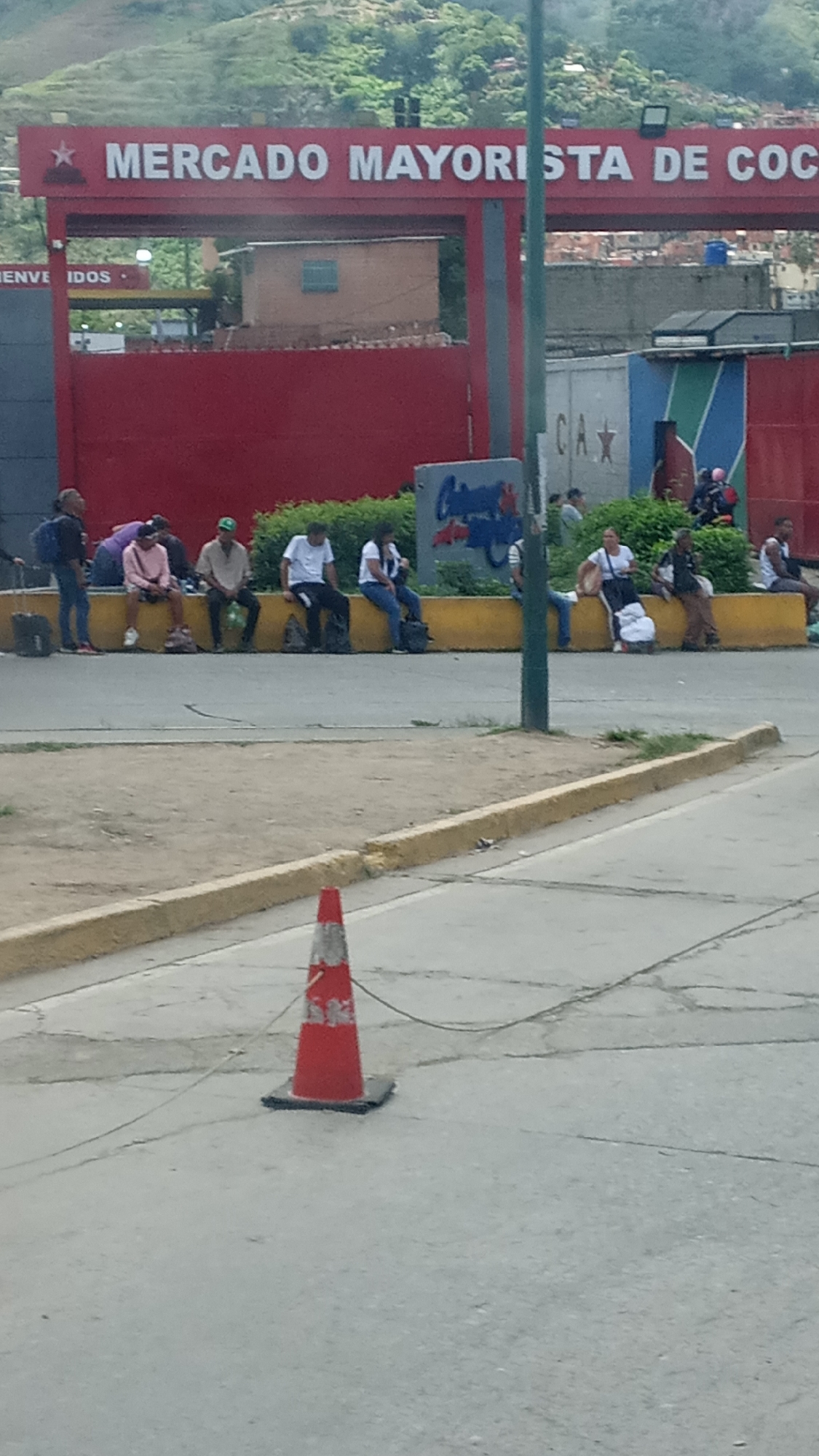 The image shows a group of people sitting on a concrete barrier in front of a red archway that reads "MERCADO MAYORISTA DE COCO," indicating a wholesale coconut market. An orange traffic cone is in the foreground. The location appears to be urban, near a hillside, with various structures and buildings visible in the background. The scene is outdoors, likely a public space adjacent to the market.