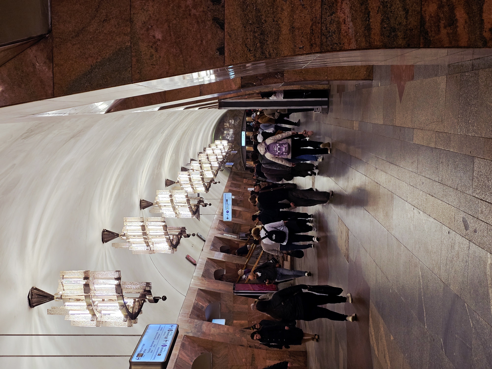 This image shows the Mayakovskaya metro station in Moscow, Russia. The station has a high arched ceiling, adorned with multiple illuminated chandeliers, and walls lined with reddish-brown marble. A line of people is visible, some are walking, and some are waiting. Signs with arrows and text are also visible along the wall, indicating directions and information. The floor is made of gray stone tiles.