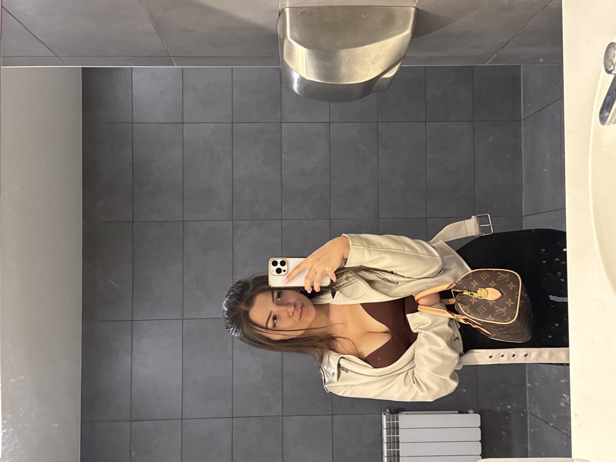 A young woman stands in a bathroom, taking a selfie with her smartphone. She is wearing a beige leather jacket over a brown top, and a black skirt or pants. A brown designer handbag rests near her hip. The bathroom features dark gray tiled walls and a white sink with a chrome faucet. A silver hand dryer is mounted on the wall. A white radiator is visible near the bottom of the frame. The image was taken in Sofia, Bulgaria.