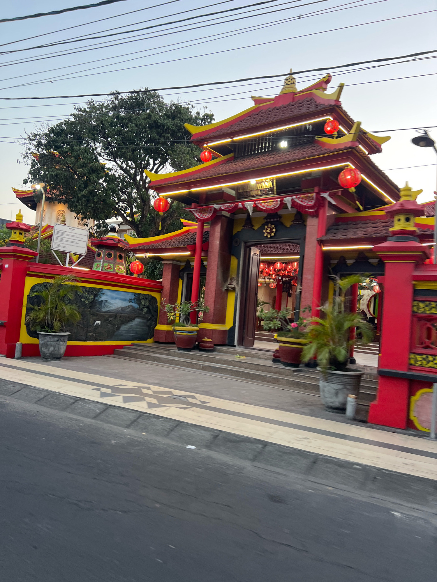 The image depicts a vibrant temple entrance in Kuta, Indonesia, at dusk. The temple features a traditional East Asian architectural style with red and yellow painted walls, ornate roofs, and numerous red lanterns. A large mural depicting a natural landscape adorns the wall to the left of the entrance. Several potted plants are strategically placed around the entrance, adding greenery to the scene. The street in front of the temple is paved with asphalt, and a decorative tiled walkway separates it from the temple grounds. Overhead, power lines crisscross the sky. The lighting suggests it is either early morning or late evening.