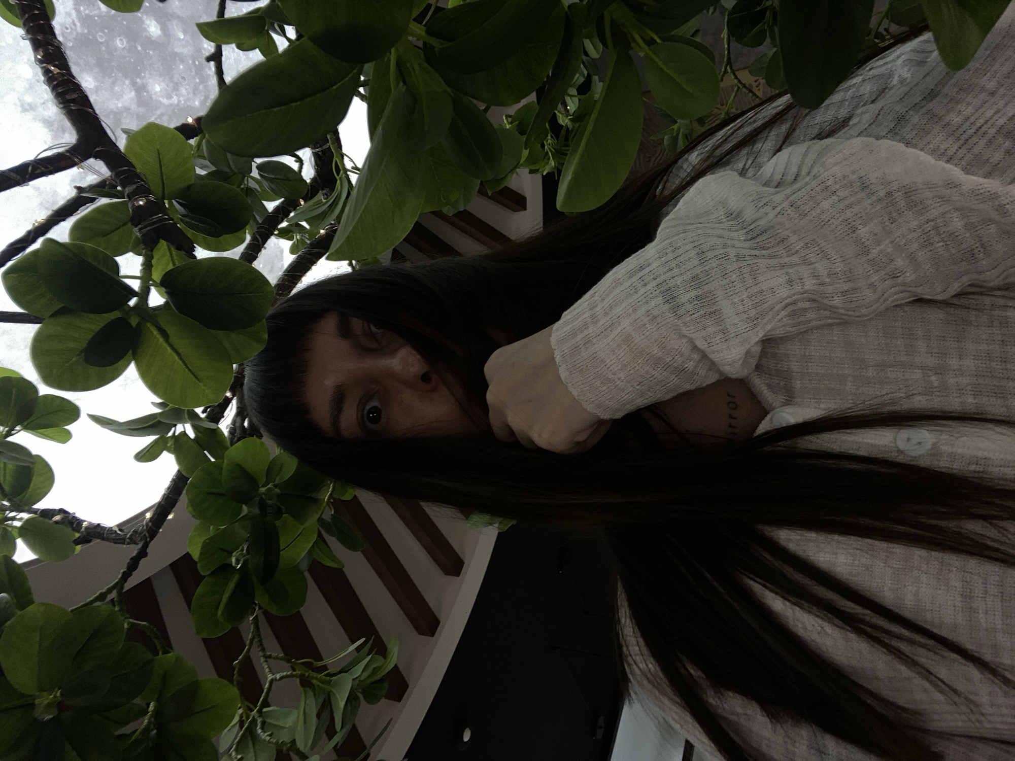 An upward-angled, candid portrait captures a person with long, dark hair, partially obscuring their face, looking directly at the camera with a wide-eyed, curious expression. Their right eye is prominently visible. They are wearing a light-colored, possibly off-white, loosely woven or textured long-sleeved top, with a button visible on the placket. Their left arm or hand is raised, contributing to the partial concealment of their face. On their neck, below the ear, a partial tattoo showing the letters "rror" is visible.
The person is positioned among lush green leaves and brown branches of what appear to be artificial plants, which are adorned with subtle decorative lights. Behind the foliage, a large, bright, circular or semicircular structure resembling a full moon with crater-like textures, or possibly a cloud-covered sky, forms the ceiling or a decorative panel. Below the plants and to the right, a patterned background of dark and light (possibly wooden) slats or beams is visible, suggesting an indoor architectural feature. The overall setting appears to be a stylized interior space, possibly a cafe or lounge, in Votkinsk, Russia. The perspective is from directly below the subject, looking up.