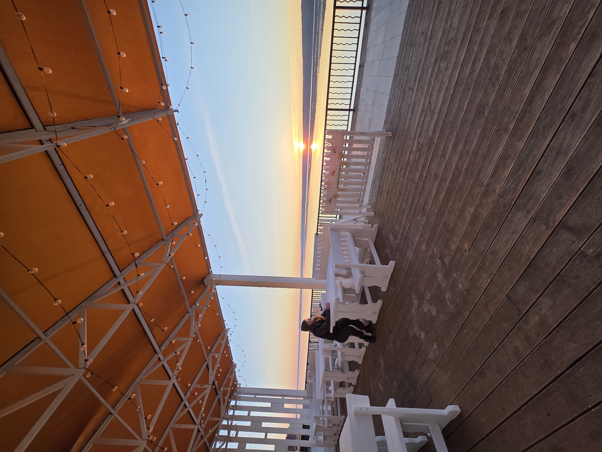 This image depicts an outdoor seating area at either sunrise or sunset, overlooking a large body of water, likely in Chaikovsky, Russia. The sky showcases a striking gradient of blue, soft orange, and yellow, with the sun visible as a bright point on the horizon, casting a luminous reflection across the calm water.
The setting features a spacious deck made of dark wooden planks. To the left, a large orange canopy, supported by a white metal framework, extends overhead, strung with numerous small, round lights. Several white wooden picnic-style tables and benches are arranged on the deck, providing seating for visitors.
One person, dressed in dark clothing and a hat, is seated alone at one of these white tables. They are facing the expansive water and the horizon, appearing to be in a contemplative posture with their head resting on their hands, possibly looking down or simply taking in the view. A dark, decorative metal railing separates the deck from the water.
The time of day is clearly marked by the warm, golden light of the sun low on the horizon, creating a peaceful and picturesque atmosphere. There is no visible text within the image.
