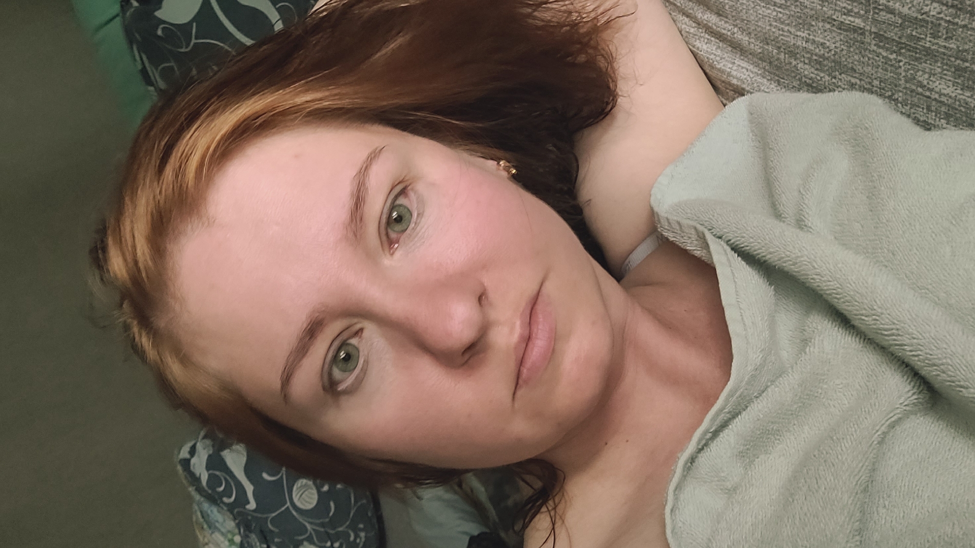 A woman with auburn hair and green eyes is lying down, facing the camera. She is covered from the chest down with a light green fuzzy blanket. Her left arm is bent and resting behind her head, while her right arm is bent in front of her chest, also covered by the blanket. She is wearing a white bra. The background appears to be a bed with patterned pillows and a textured grey headboard. The lighting is dim, suggesting an indoor setting, possibly a bedroom. The scene is static and intimate, capturing a candid moment.