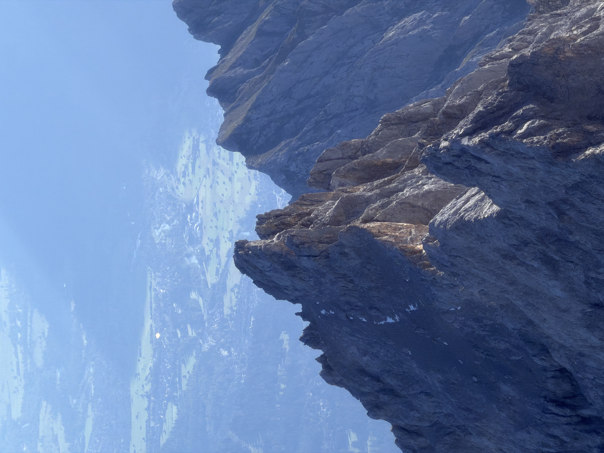 This is a photograph taken from a high vantage point, likely a mountain peak, looking down upon a valley and a town. The foreground is dominated by rugged, grey, rocky mountain ridges with sharp peaks and steep inclines. There are patches of snow on some of the rocky surfaces.
In the middle ground, the terrain transitions to a green valley floor dotted with trees and structures of a town. The town appears to be nestled amongst hills and meadows. A notable feature in the town is a bright, circular object, possibly illuminated by sunlight reflecting off a dome or a similarly reflective structure. Roads or pathways are visible winding through the landscape.
The background consists of a hazy blue sky and a distant, blurred landscape, suggesting a significant amount of atmospheric haze or distance. The overall impression is one of vastness and natural grandeur, with human habitation appearing small and integrated into the mountainous environment. The location is specified as Grainau, Germany, a town known for its proximity to the Alps.
