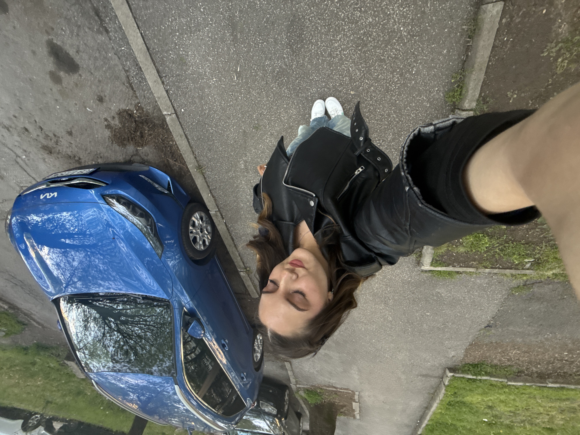 A tilted, wide-angle, full-length photograph depicts a young woman standing on an asphalt surface next to a blue Kia sedan. The woman has brown hair and is wearing a black leather jacket, light blue jeans, and white sneakers. She appears to be holding the camera herself, with an arm and portion of a black-clad shoulder visible in the foreground. The car is parked near the curb, with the front of the car facing the camera. The asphalt surface shows signs of wear and contains dirt and minor vegetation along the edges, where it meets with concrete curbs and small patches of grass. Reflected on the car's windshield, one can see tree branches and the sky. The surroundings suggest an urban area, potentially a residential street. Given the architecture, the picture was taken in the city of Sofia, Bulgaria.