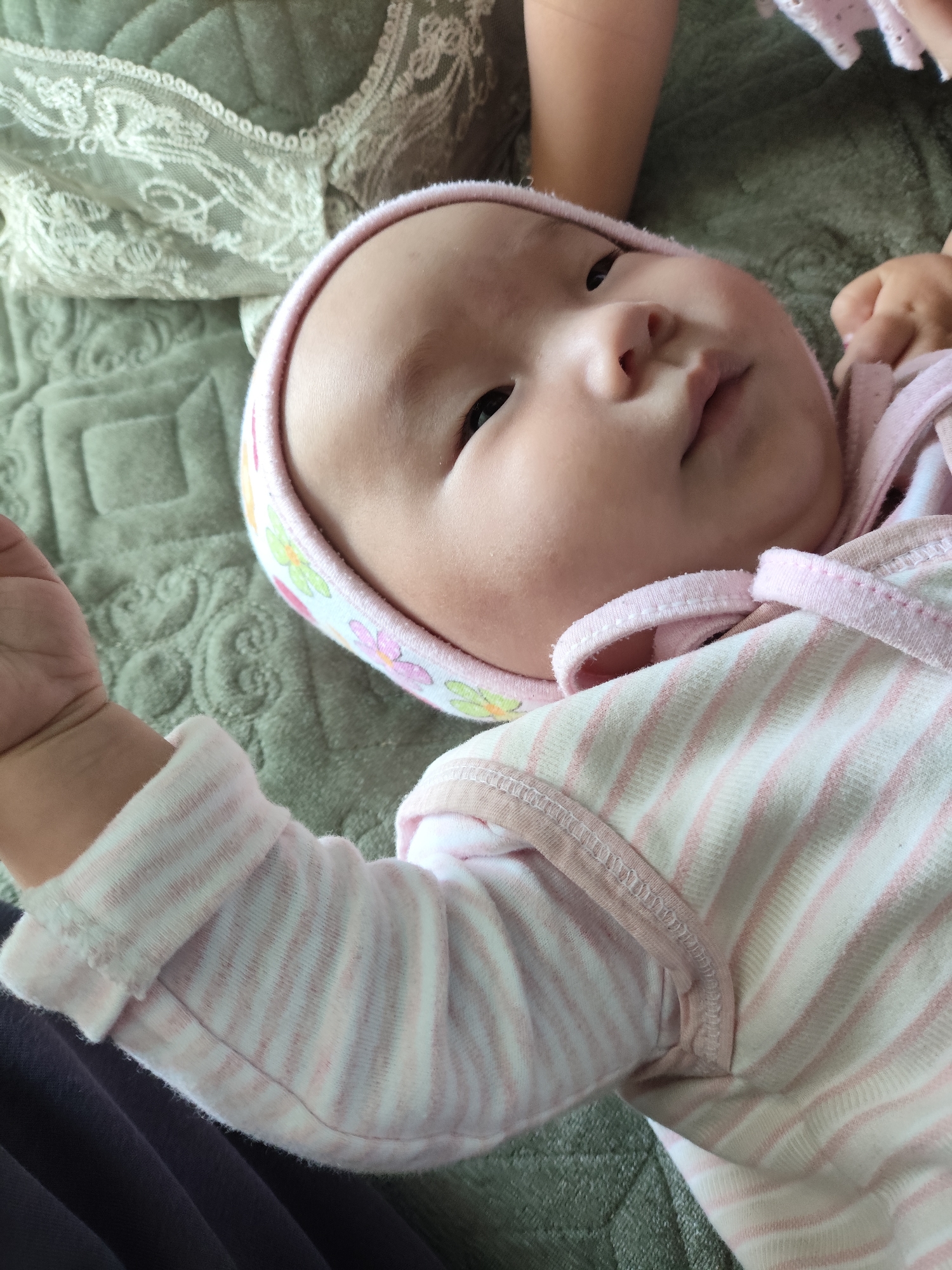 An overhead, close-up, eye-level, medium shot captures a baby lying on a quilted, grey blanket. The baby, facing upwards, wears a pink and white striped onesie and a matching hat decorated with small colorful flowers. The baby's face, with its eyes closed and mouth slightly open, is partially obscured by the hat. The baby's left arm is extended outwards, with its hand visible in the lower left corner of the frame. The baby is positioned in the center of the frame, with a portion of another person's arm visible in the upper right corner. The overall scene suggests a peaceful moment of rest or play. The image does not contain any discernible elements that would allow for the identification of its location within the city of Tasma, Kyrgyzstan.