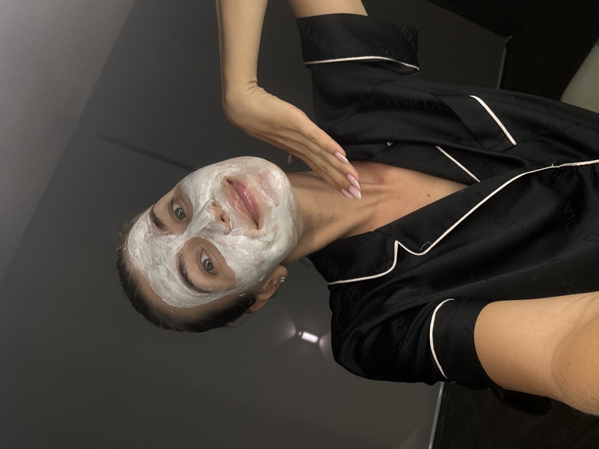 A woman is pictured with a white facial mask. She is wearing a black silk pajama top with white piping. Her manicured hand is resting on her neck. The location is indoors.