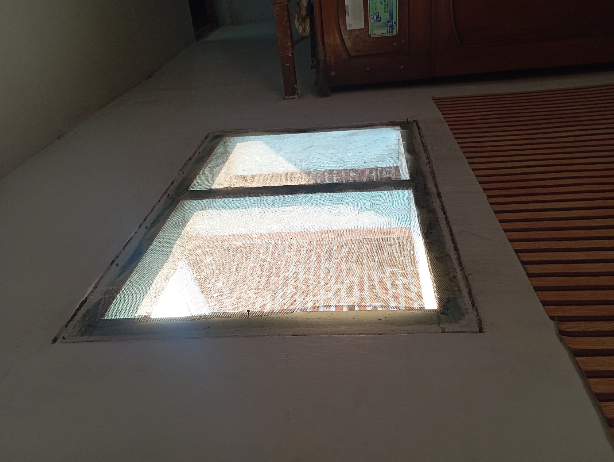 The image shows a close-up view of a window from the inside of a room. The window has a double pane and is covered with a fine mesh screen. Outside the window, a brick wall is visible, illuminated by sunlight. The room appears to be sparsely furnished. To the right of the window, a wooden door with decorative carvings is partially visible. The overall condition of the room suggests it may be older or in need of some repair, with dust and wear visible on the window frame and walls. The lighting suggests it is daytime. This scene is consistent with a residential interior in Jogosari, Indonesia, as indicated by the style of the window, door, and the general aesthetic which aligns with common housing in the region.