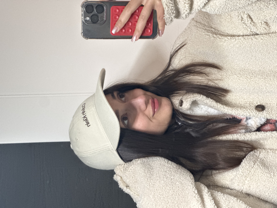 The image captures a close-up, sideways mirror selfie of a young woman. Her head is positioned on the left side of the frame, with her body extending to the right.
The woman has long, dark brown hair that flows around her face and over her shoulders. She is looking slightly upwards and to her left (from the viewer's perspective), with a subtle, relaxed smile on her lips, which appear glossy. She is wearing a light-colored (beige or cream) baseball cap, with the brim facing away from the camera.
She is dressed in a very fluffy, cream or beige-colored jacket, likely made of sherpa or fleece material, with several brown buttons visible down the front. A glimpse of a patterned garment, possibly in shades of red and pink, can be seen underneath her jacket near her neck.
Her right hand is visible in the upper part of the reflection, holding a smartphone. The phone appears to be an iPhone with multiple camera lenses, encased in a clear case with dark edges. Attached to the back of the phone case is a red, rectangular grip with circular indentations, resembling a pop-it fidget toy. Her fingernails are short and styled with a white tip, indicative of a French manicure.
The background is simple and appears to be an indoor setting. It consists mainly of a plain, light-colored (white or off-white) wall, with a darker section (possibly dark grey or black) visible on the lower left side, creating a two-toned effect or indicating furniture.
The visible text on the baseball cap reads "nieLK nivlaC," which is the reversed and upside-down reflection of the brand name "Calvin Klein."
