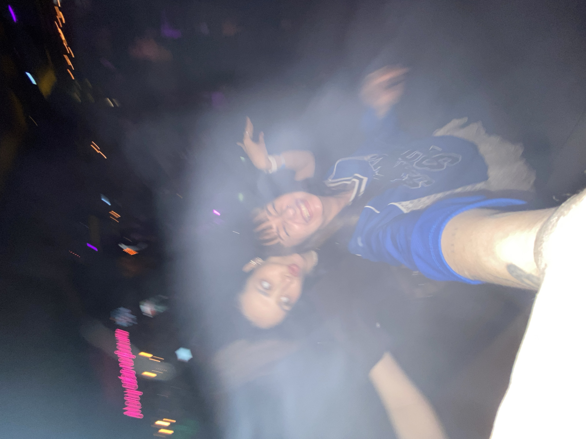 This blurry and rotated image captures two young women in a dimly lit, smoky environment, likely a concert, club, or party. The primary light source appears to be a camera flash, causing significant reflection in one woman's eyes.
The image is heavily rotated clockwise. When oriented upright, the woman on the right is smiling broadly at the camera, with her left arm raised, possibly waving or dancing. She has dark hair with bangs and is wearing a blue and white top that resembles a varsity jacket or sports jersey, with some white text or a logo on the chest that is unreadable due to blur.
Below her and slightly to the left, the second woman looks directly at the camera with a more neutral expression. Her dark hair frames her face, and her eyes reflect the flash intensely, appearing bright white. She is wearing dark clothing.
The background is very dark, punctuated by various streaks and blurs of colored lights – pink, orange, purple, and blue – indicating artificial illumination typical of a live event venue. A significant amount of smoke or fog diffuses the light and contributes to the overall blurry effect. On the far left, a vertical line of bright pink, wavy lights, possibly decorative or stylized unreadable text, is visible. The overall mood conveyed is energetic and lively, capturing a moment of enjoyment during a nighttime event.