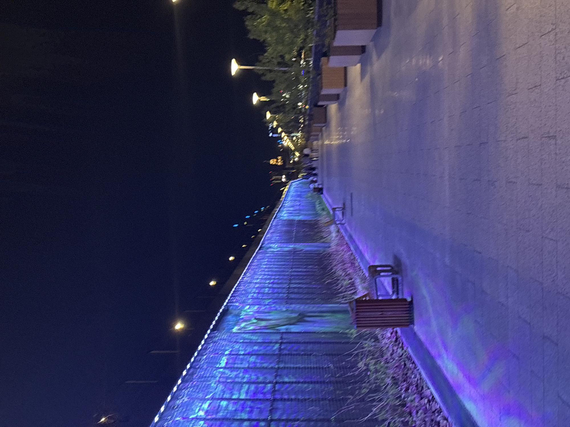 The image depicts a nighttime scene along the Kama River embankment in Perm, Russia. The embankment is illuminated by numerous streetlights, casting a glow over the pedestrian walkway and the river's edge. The walkway is paved with stone and features wooden benches placed along its length. To the left, the river is visible, with its surface reflecting the city lights in blue hues. In the background, the city skyline is visible, with buildings illuminated in warm colors. The sky is dark, adding to the nighttime atmosphere.