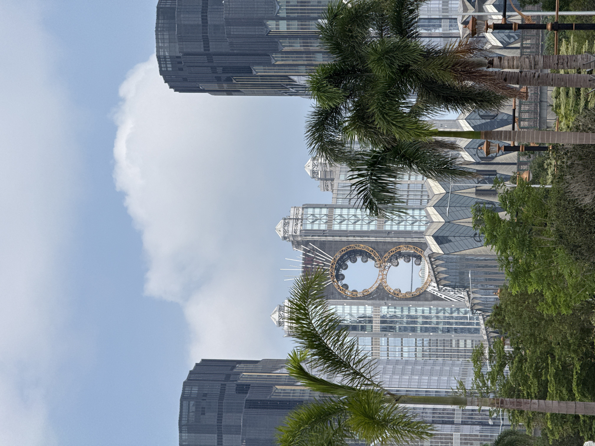 The image depicts a cityscape with a cloudy sky above. In the center is a building with a distinct architectural feature consisting of two large, interconnected Ferris wheels. Palm trees are positioned in the foreground, partially obscuring the view of the buildings. The buildings are modern, with glass and steel structures. Vegetation, including trees and shrubs, lines the base of the buildings. The scene is brightly lit, suggesting a daytime setting. The location is likely in the city of Unknown, Macao.