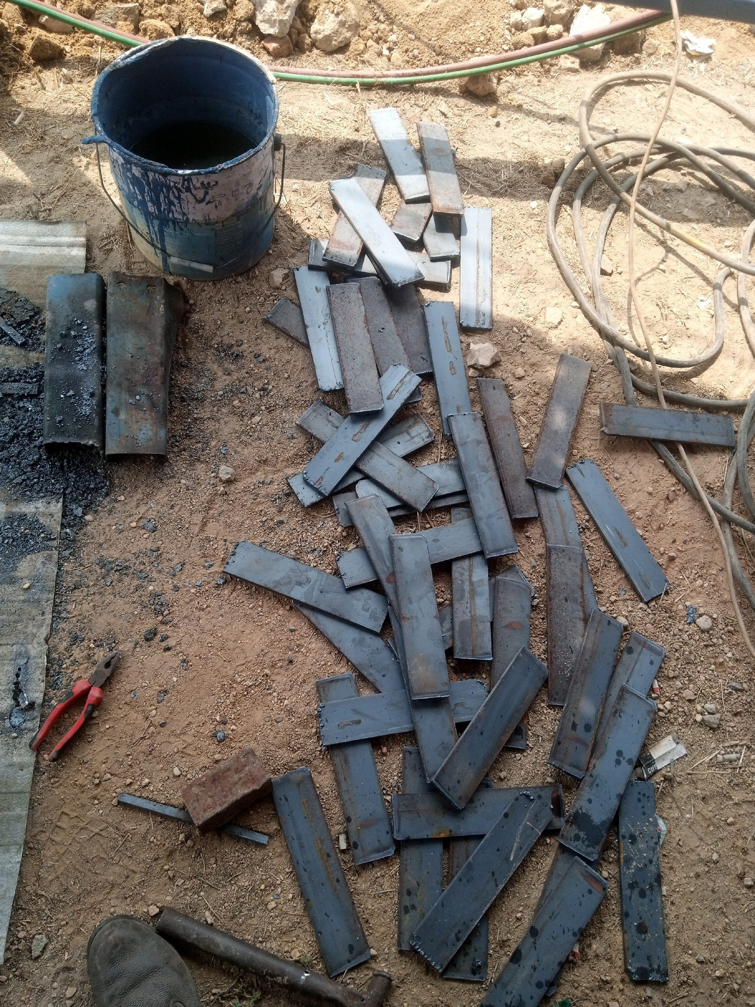 This image depicts a construction or workshop scene on the ground in Teluk Dalem Ilir, Indonesia. The foreground shows a worn boot, a brick, a rusty metal bar, and red-handled wire cutters. Scattered across the sandy ground are numerous metal rectangular pieces, likely scrap metal or materials for fabrication. A blue metal bucket, containing a dark liquid and showing signs of wear and paint drips, is also present. In the background, a thick bundle of dark electrical cables or hoses lies on the dirt, along with some rocks and debris, suggesting an outdoor work environment. The lighting indicates it is daytime. No people are visible in the image.