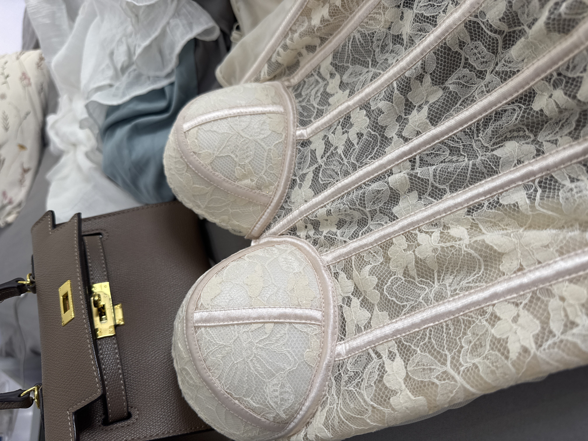 The image depicts a close-up view of a light beige lace corset lying next to a dark beige leather handbag. The corset features structured cups with light beige trim, a lace overlay, and boning details. The handbag is partially visible, showcasing its gold-toned hardware and structured design. Both items are placed on a soft, gray surface, likely a bed or similar cushioned area. Additional soft fabrics, including white and blue textiles with ruffles, are visible in the background, lending a cluttered yet casual atmosphere. The location is impossible to determine based on the visible information.