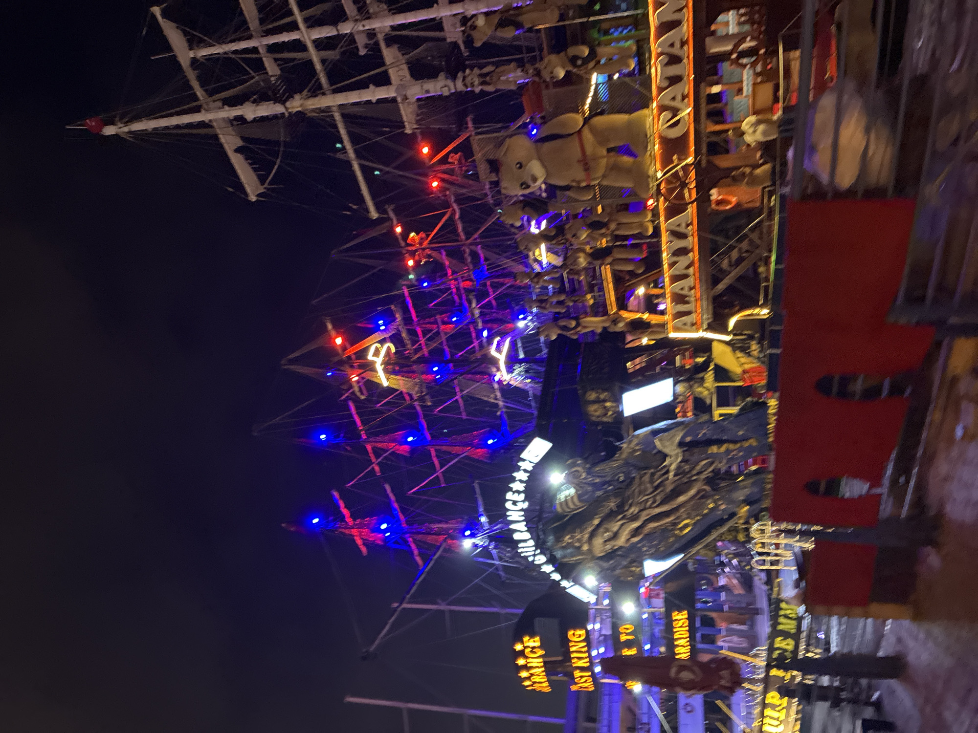 The image captures a nighttime scene in Alanya, Turkey, featuring a large, elaborately decorated ship, likely a pirate-themed attraction or vessel. The ship is illuminated with numerous bright lights, primarily in red and blue, accentuating its masts, rigging, and hull. Various signs are visible on and around the ship, including "ALANYA CATAMARAN" and other text that is partially obscured or illegible due to the lighting and angle. Several teddy bear figures are displayed on the upper deck of the ship, adding to its festive or whimsical appearance. The foreground shows a red barrier or fence, suggesting a viewing area or pathway. The overall atmosphere is lively and festive, indicative of a tourist area at night.