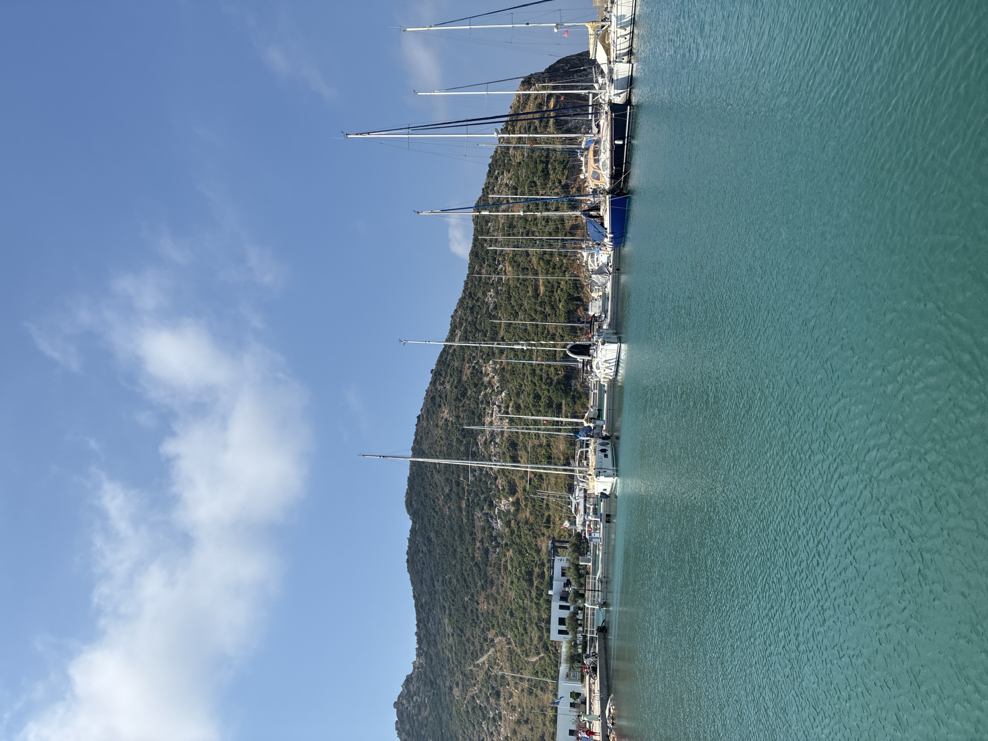 A wide-angle, eye-level outdoor shot depicts a marina with numerous sailboats docked. The water is a clear, turquoise color with small ripples. In the background, a green, rocky hill rises behind a row of white buildings. The sky is a clear blue with a few wispy white clouds. The sailboats are various sizes and colors, with white hulls predominating. Several people are visible on the dock, walking and standing. The scene appears to be a sunny day in Gazipaşa, Turkey.