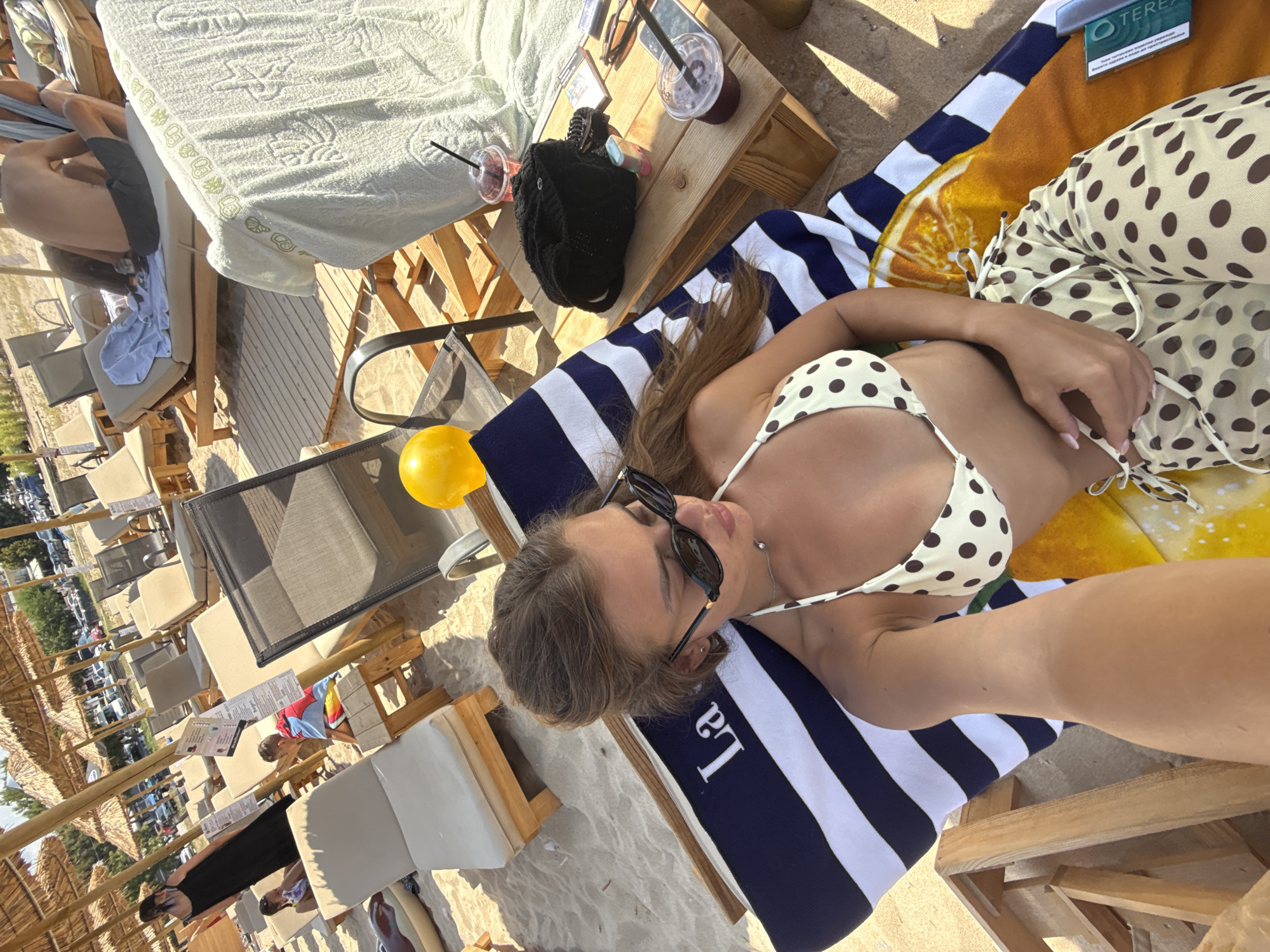 The image captures a scene at Ofrynio Beach, Greece, under a bright, sunny sky. In the foreground, a young woman lies on a beach chair, wearing a polka dot bikini and matching pants. She is captured in a selfie-style shot, with her head and upper body prominently featured. She wears dark sunglasses and has her hair pulled back. Beside her, a striped towel with the word "La" visible is spread across the beach chair.
The midground reveals more of the beach setting with rows of empty loungers and umbrellas, suggesting a popular tourist destination. A small wooden table is placed near the woman's chair, holding a black bag, two drinks in clear cups with straws, sunglasses, and a phone. In the background, other beachgoers can be seen relaxing on loungers, and parked cars are visible beyond the beach area, indicating proximity to a road or parking lot. The overall atmosphere is one of leisure and summer vacation.