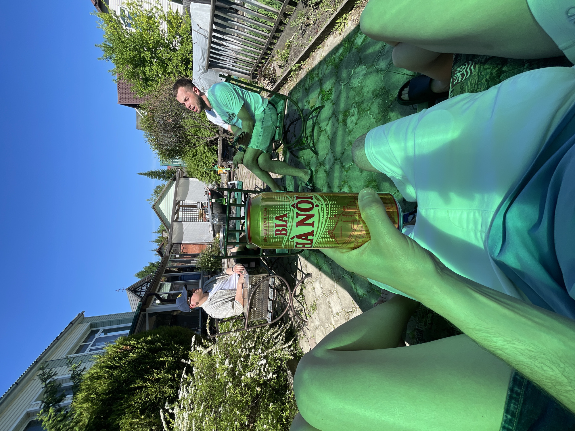 The image captures an outdoor scene in a backyard or garden in Манский, Russia, under a clear blue sky and strong sunlight. The perspective is first-person, showing the legs and arm of an individual wearing white shorts and holding a golden "BIA HÀ NỘI" beer can.
Two other men are present, seated on metal garden chairs. To the left, a man wearing a blue baseball cap, sunglasses, and a light plaid shirt over a white t-shirt sits at a small round table, looking to his right. A metal chest-like object, possibly a grill or cooler, is positioned near his chair. Further in the background, a house with light green siding and a white trim is visible.
To the right, a bearded man wearing a two-tone green and white t-shirt, light denim shorts, and dark flip-flops, leans back in his chair with his eyes closed or squinting and mouth slightly open, appearing to relax in the sun. A smartwatch is visible on his left wrist. A wooden fence and more residential buildings are in the background on the right.
The ground comprises stone paving and a prominent green ground cover or tarp, which imparts a strong green tint to the lower half of the image. Lush green bushes, including a flowering white shrub, are visible throughout the setting.