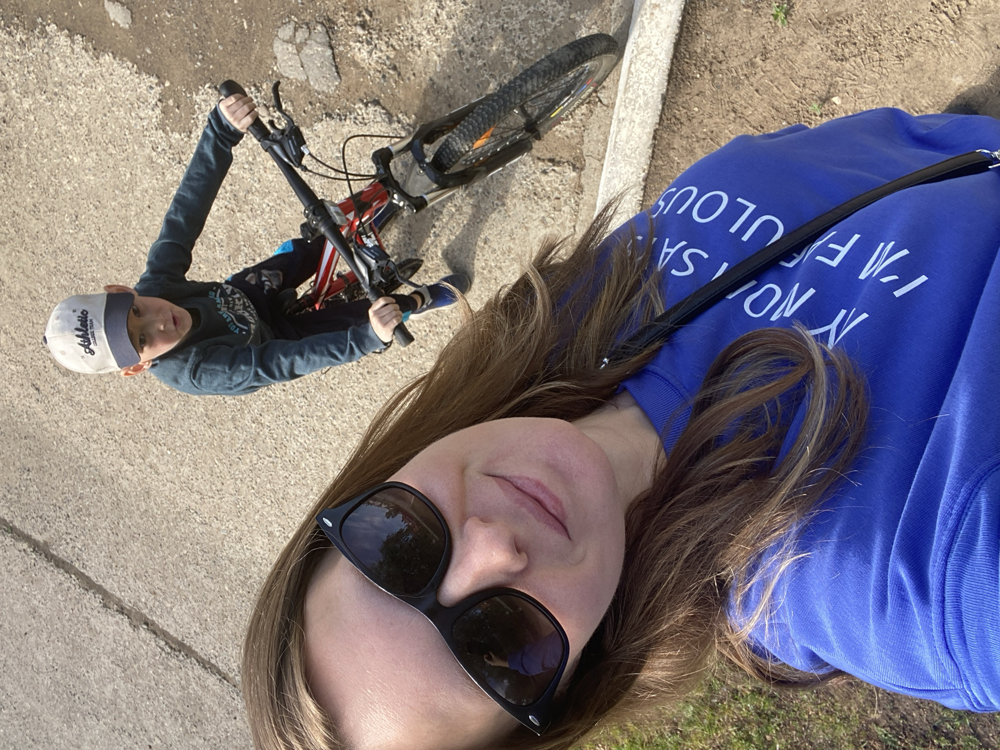 A selfie is taken from an elevated perspective, capturing two individuals. The person taking the photo has fair skin, sunglasses, and brown hair. They are wearing a royal blue sweatshirt with white text that reads, "I'M NOT SAYING I'M FABULOUS". A black strap is also visible, likely from a bag or purse.
To the left of the photographer, a child is shown riding a bicycle. The child is wearing a hat and a long-sleeved shirt, and is actively holding onto the bicycle handlebars. The bicycle is primarily red and black.
The setting appears to be outdoors on a paved surface, possibly a sidewalk. A concrete curb is visible near the top right of the frame. The location is Noviy, Russia.