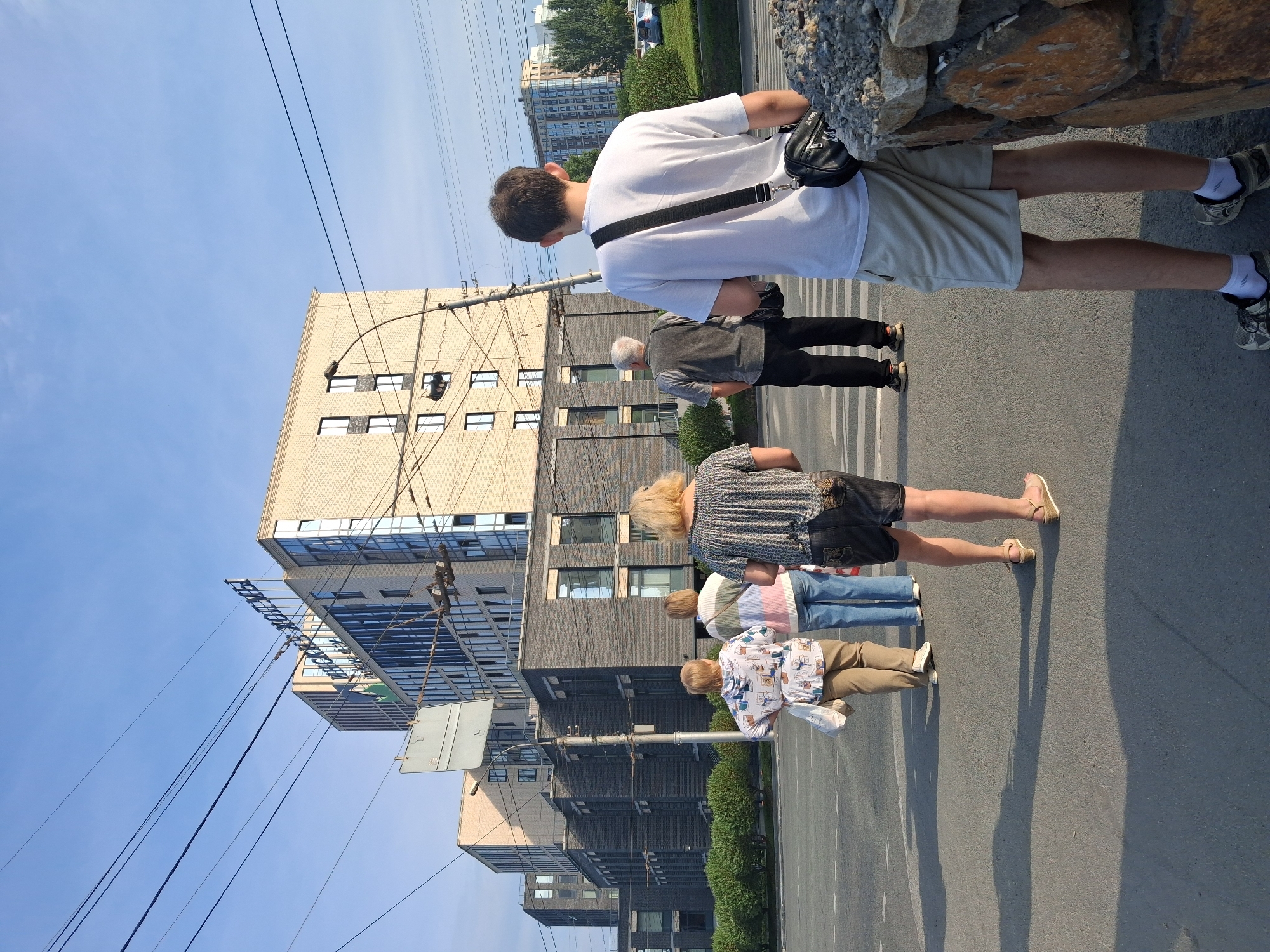 In Novosibirsk, Russia, on a clear day, a group of five individuals is observed standing on a crosswalk in front of a modern multi-story building complex. The building features a combination of light-colored brick and dark brick facades, with numerous windows. A large sign with blue lettering reading "АНКОМ" is visible on the top of one section of the building. Overhead power lines crisscross the sky.
From right to left, the individuals are:
1. A young male, seen from the back, wearing a white t-shirt, light-colored shorts, white socks, and athletic shoes. He has a black shoulder bag slung across his chest.
2. An older male, also seen from the back, wearing a dark vest over a grey shirt and black pants, with black shoes. He carries a black bag over his left shoulder.
3. A woman with blonde hair, facing away from the camera, wearing a patterned dark top and dark denim shorts, with sandals.
4. A woman wearing a striped sweater in shades of pink, white, and blue, and blue jeans, with white shoes.
5. A woman with short blonde hair, wearing a light-colored patterned hoodie over tan pants, and light-colored shoes. She has a white bag slung over her left shoulder.
The group appears to be waiting to cross the street. The environment suggests an urban setting with paved roads, a sidewalk or median, and some landscaping with green bushes. The lighting indicates it is likely late afternoon or early morning due to the long shadows.
