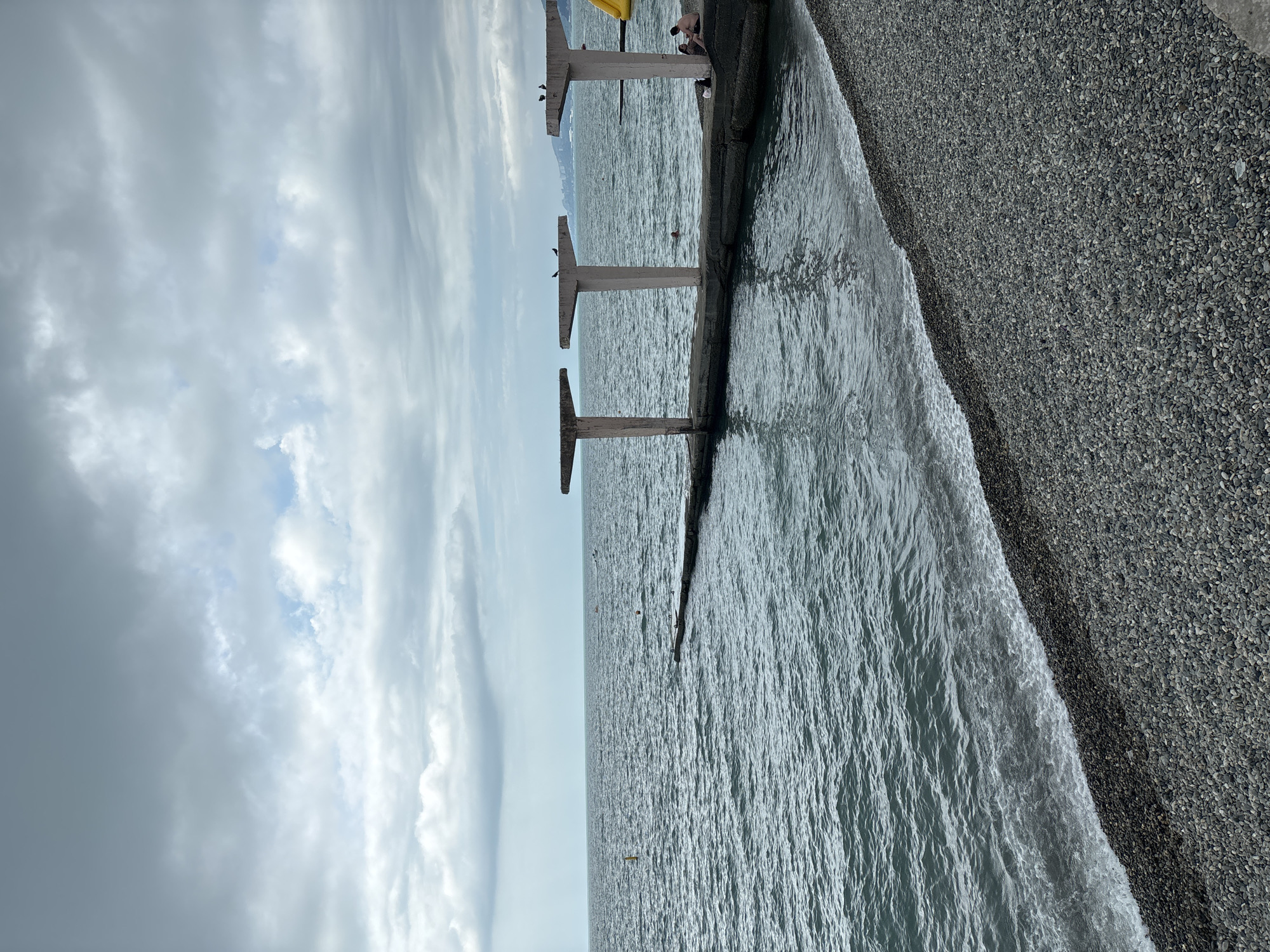 The image shows a cloudy day at the beach in Sochi, Russia. A concrete pier with three umbrella-shaped structures extends into the sea. There are a few people on the pier and in the water, and some pigeons are perched on the structures. The beach is covered in pebbles, and the sea is choppy. The overall impression is one of a somewhat bleak but peaceful day at the seaside.