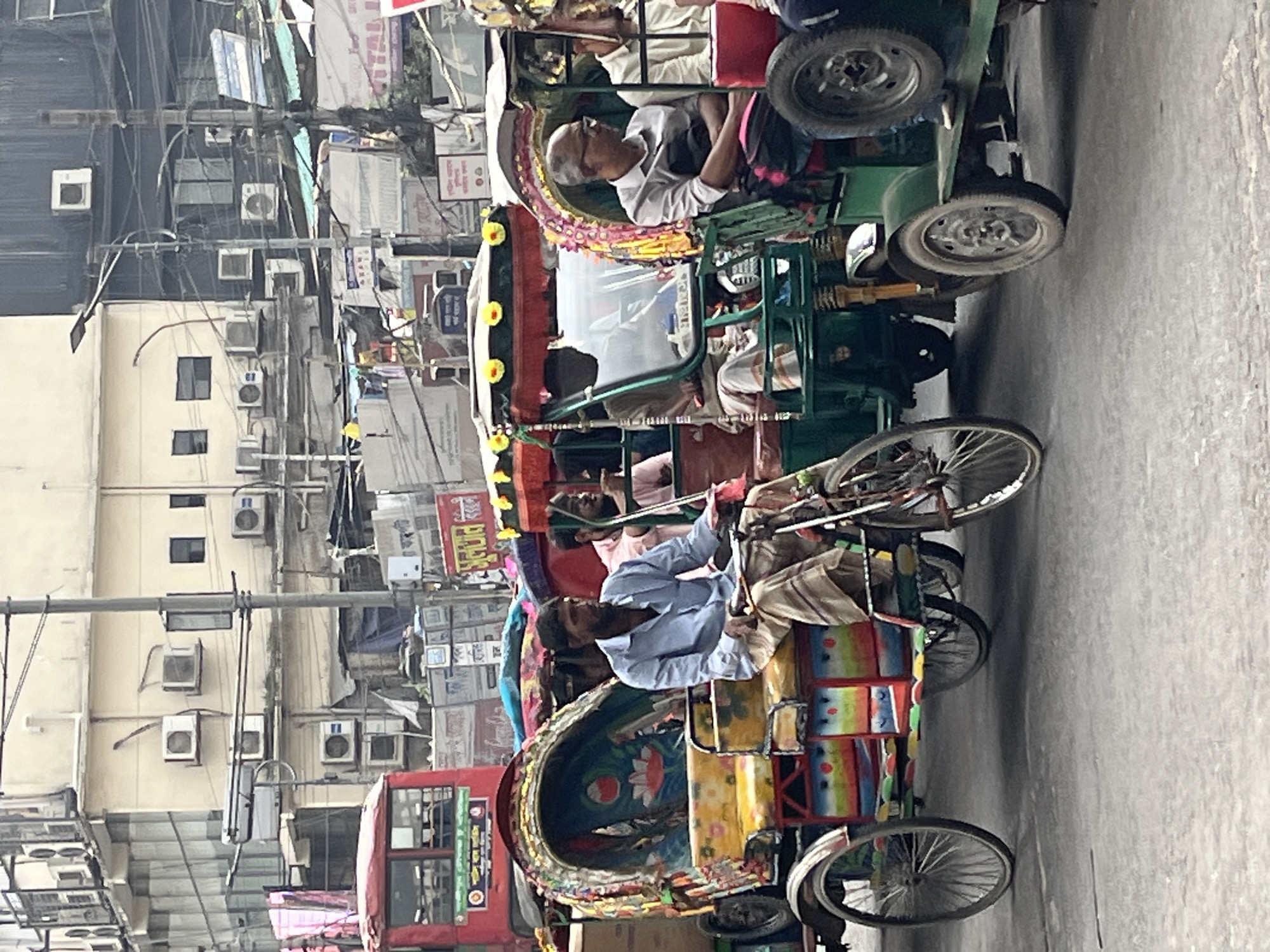 The image shows a street scene in Dhaka, Bangladesh. Multiple cycle rickshaws are in the foreground with their drivers. Each rickshaw is ornately decorated with colorful paint. Buildings with multiple air conditioning units line the background. Power lines run overhead in a tangled network. A red bus is visible further down the street. The setting appears to be a busy urban area with a high population density.
