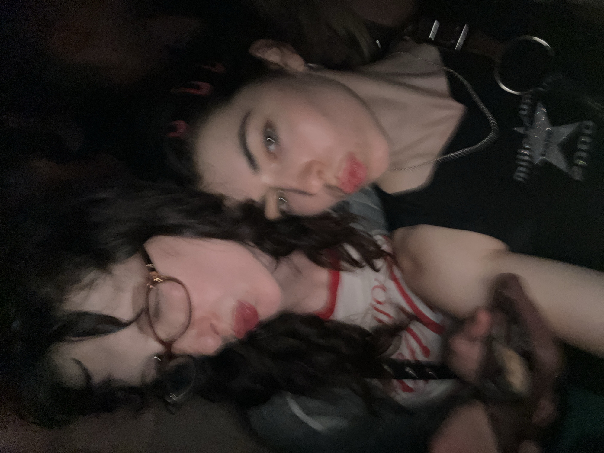 Two young women are lying close together in a dimly lit indoor environment. One woman is wearing glasses and a white and red striped shirt. The other woman has a nose ring and is wearing a black top with a star graphic. The scene appears to be candid, captured in low light conditions. The specific location in London, United Kingdom is indiscernible.