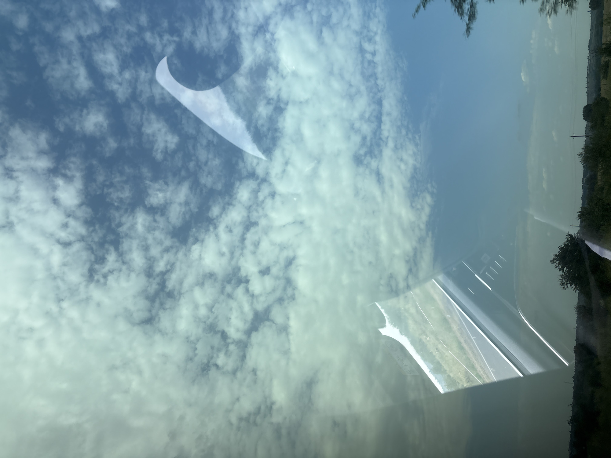 The image depicts a view through a reflective surface, likely a window, offering a distorted perspective of a cloudy blue sky and a landscape below. Several light-colored, curved reflections, possibly from the window frame or a structure, are prominent against the sky. The lower portion of the image reveals a scene with a road, green fields, and trees. The overall impression is a layered composition created by reflections on a transparent surface, superimposed over a natural environment. The mention of Vannovskoye, Russia, suggests a specific geographical context for this scene, implying that the view is from within or overlooking this location.