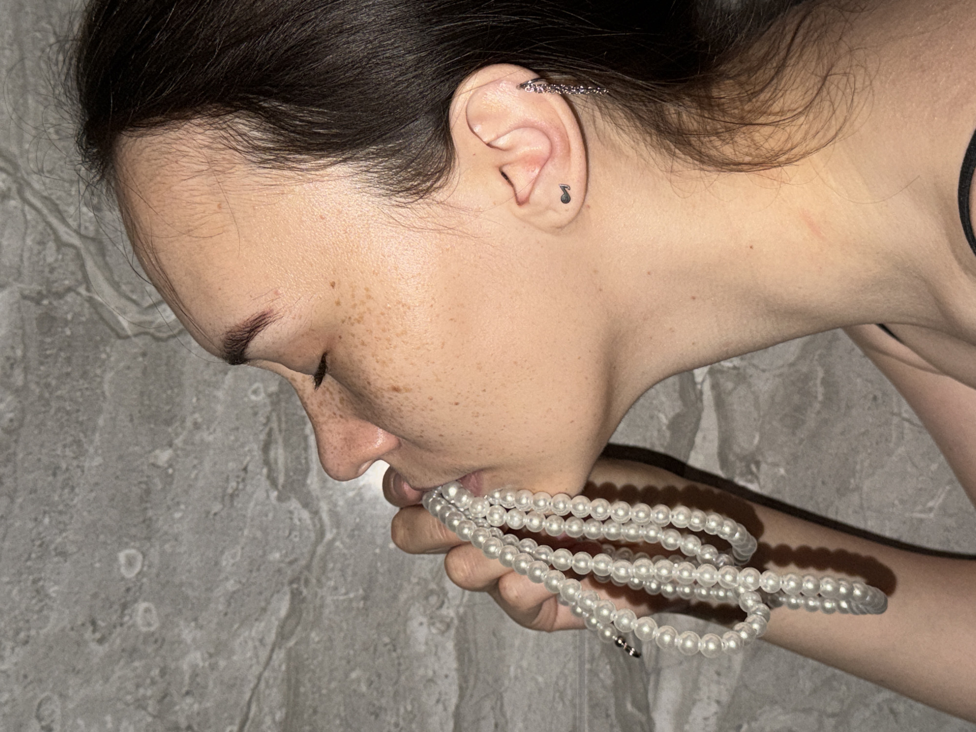 A young woman with brown hair and freckles is holding a strand of white pearls up to her mouth in a profile view. She is wearing a black strap top and has a small musical note tattoo on her earlobe and a dangling earring in her right ear. The background is a textured gray wall. The image appears to be taken indoors. There is no indication of the city of Chaikovsky, Russia in the image.