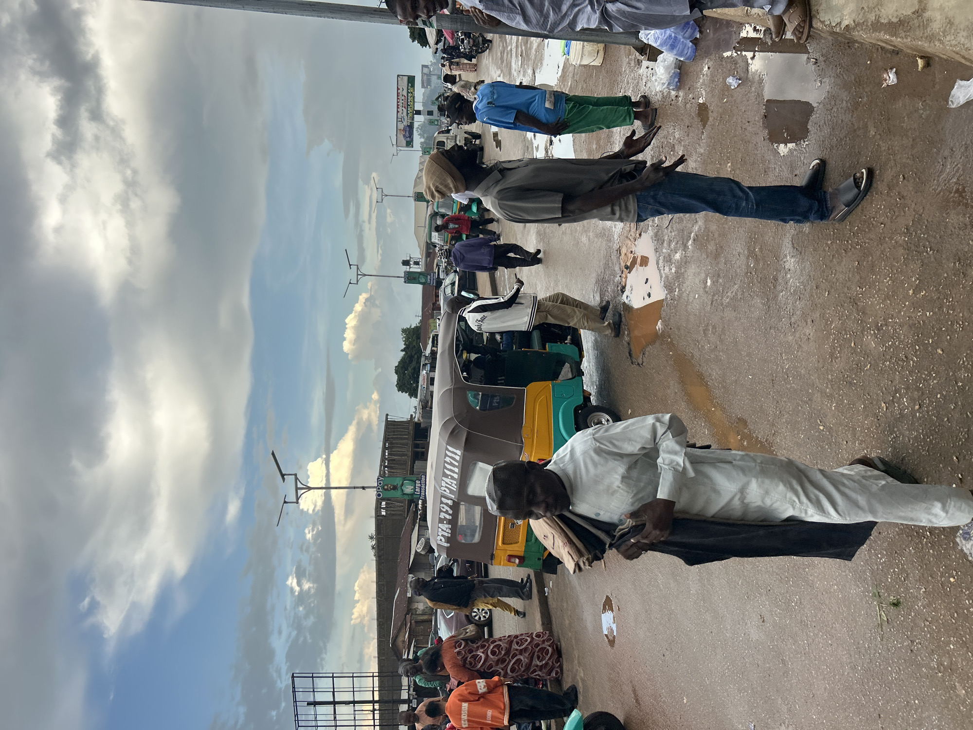 This is a street scene in Jos, Nigeria, likely during or shortly after rainfall, as evidenced by the puddles and wet asphalt. The sky is partly cloudy with dramatic, grey formations suggesting recent or impending weather changes. The environment appears to be a bustling urban area with a mix of commercial activity and transportation.
In the foreground, a man in a light-colored traditional tunic and trousers, wearing a cap, walks towards the viewer, carrying folded dark fabric. His gaze is directed downwards. To his left, a yellow and green auto-rickshaw (keke napep) with a license plate that reads "PTA-791 PTA-11711" is parked. Other vehicles, including a dark sedan and a lighter-colored car, are visible behind the auto-rickshaw.
Further back, a group of people are gathered. One individual in an orange jacket with "ECMONSI" printed on it stands facing away. A woman in a patterned dress is also present. Several men are seen in various casual attire, including jeans and t-shirts, some conversing or observing. One man wears a tan beanie and gestures with his hands, while another, wearing a blue t-shirt and green trousers, stands nearby.
The background shows low-rise buildings, some with corrugated iron roofs, and overhead power lines and streetlights. A billboard with an advertisement featuring a man's portrait and the text "Opey Large Transaction" is prominently displayed. The overall impression is of a lively, everyday scene in a Nigerian city.