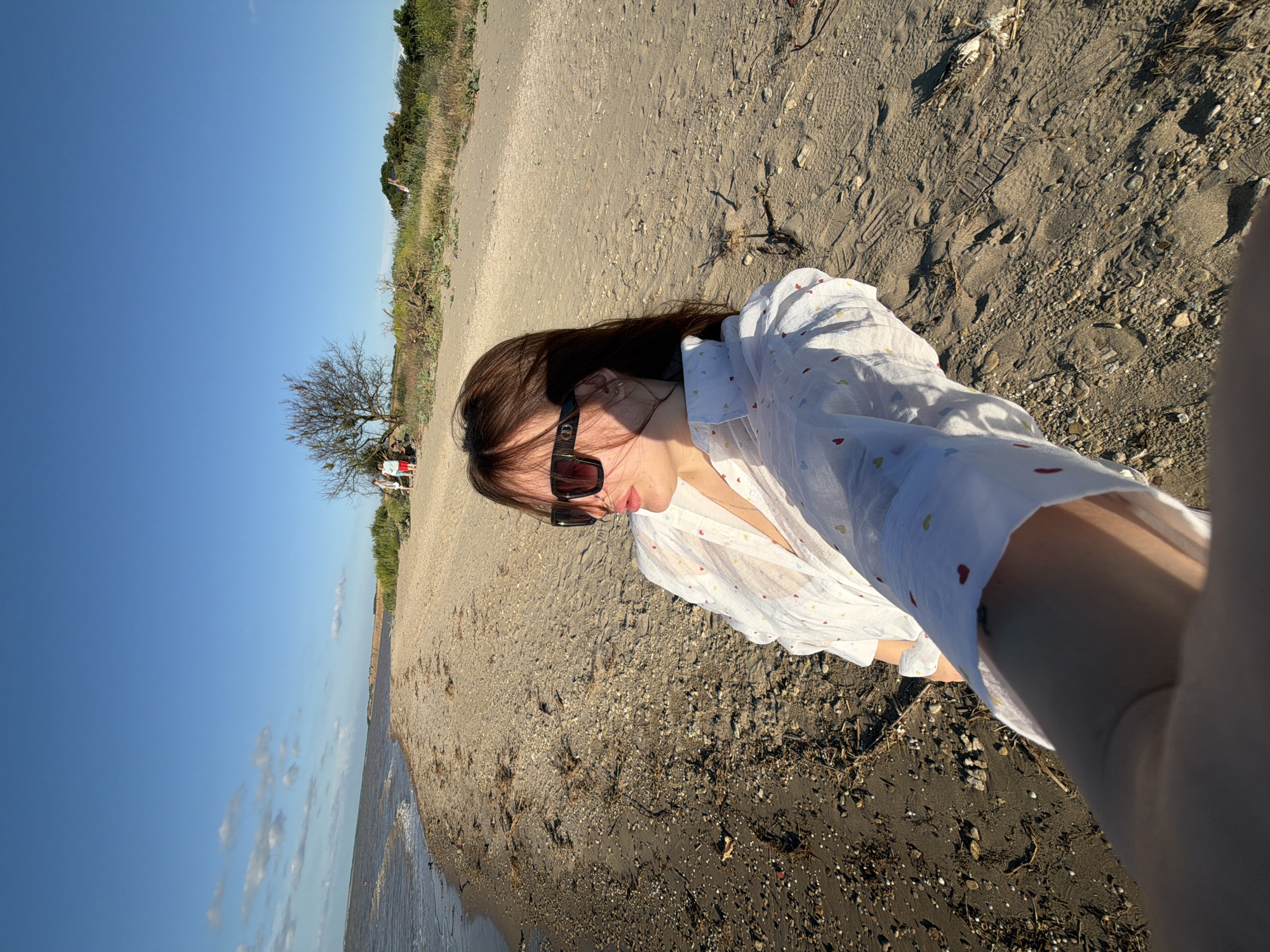 The media file depicts a selfie taken by a person on a sandy beach near Port-Katon, Russia. The subject is wearing sunglasses and a white shirt with a pattern of small red and yellow hearts. The background shows a clear blue sky, a shoreline with gentle waves, and a vegetated area with a bare tree and some shrubs. In the distance, a few other people are visible near the vegetation, some of whom appear to be seated. The sand on the beach is dark and contains small pebbles and debris. The lighting suggests it is daytime, likely late afternoon given the shadows. The overall mood appears relaxed and leisurely, consistent with a day at the beach. There is no visible text in the image.