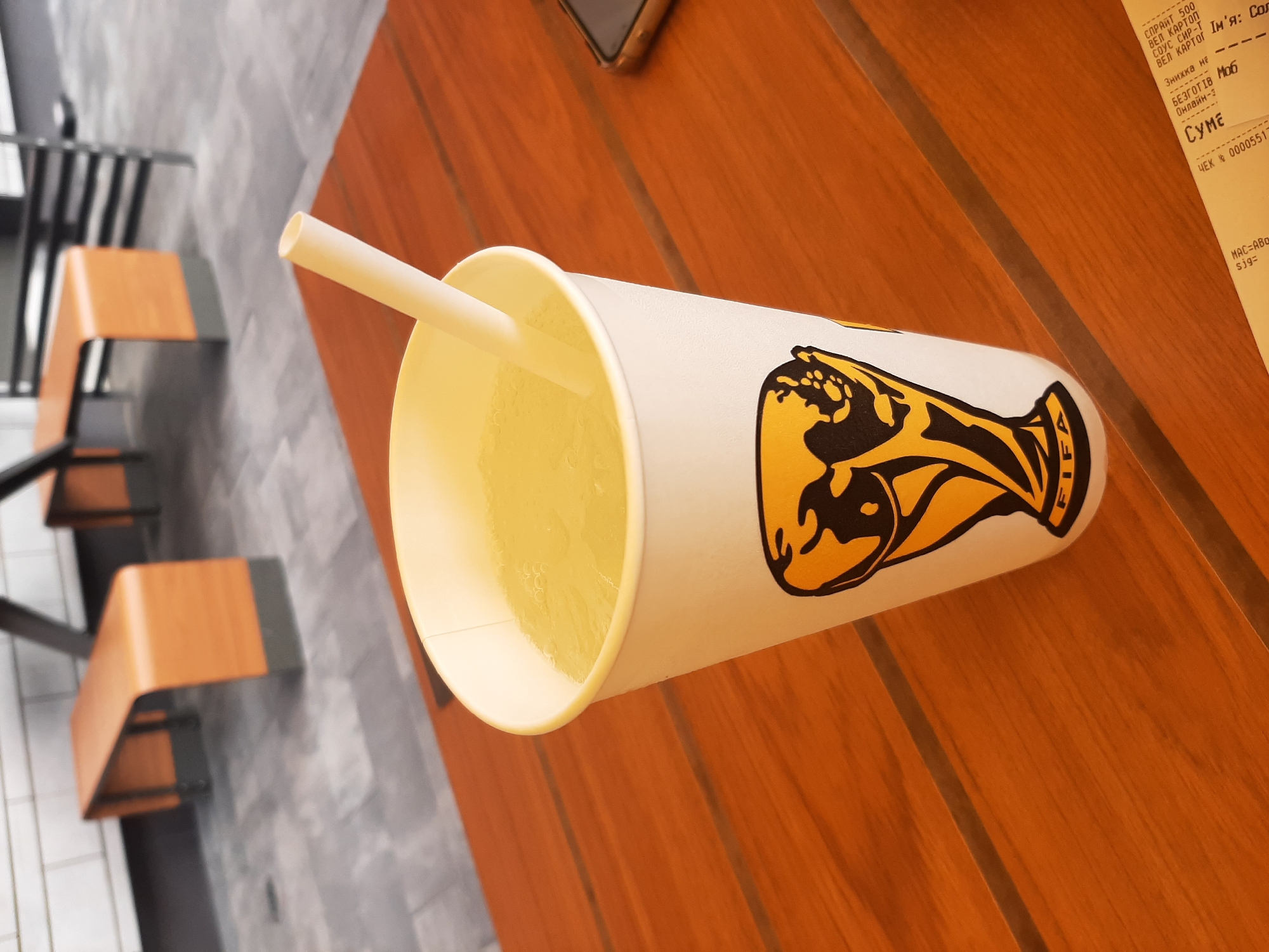 A disposable white paper cup with a straw is placed on a wooden table. The cup features a black and yellow FIFA World Cup trophy logo. The table is set outdoors, with grey tiled flooring and modern wooden benches visible in the background. A portion of a mobile phone is visible on the right side of the table. A receipt is partially visible at the bottom right corner. The overall scene suggests a casual outdoor dining or resting area, possibly in a public space like a park or a cafe with outdoor seating in Lviv, Ukraine.