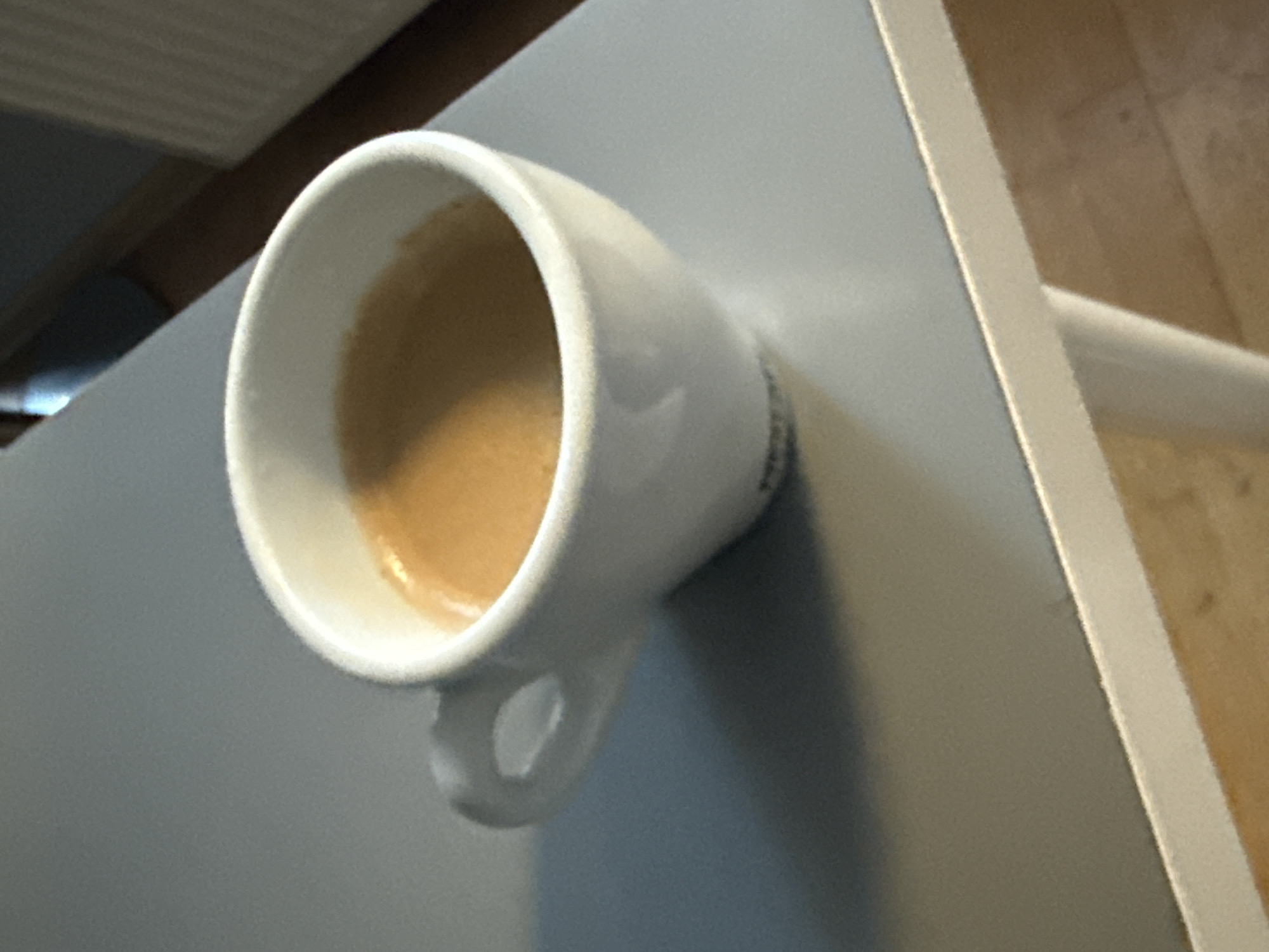 A close-up shot shows a white espresso cup filled with coffee, placed on a grey surface that appears to be a table. The coffee has a light brown crema on top. The cup has a white handle, and the overall lighting suggests an indoor setting, possibly a cafe or home. The background is blurred, with a hint of a radiator or wall in the upper right corner. The scene is devoid of any people or specific landmarks that would pinpoint its location in Wrocław, Poland, or any other city.