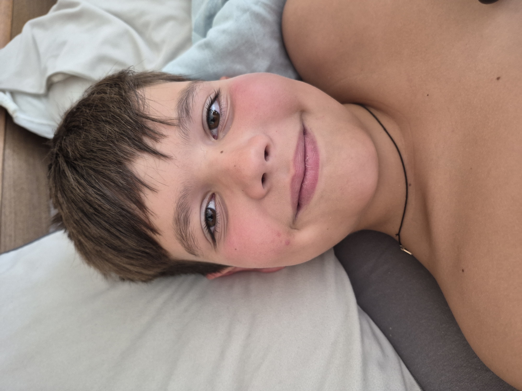 The image is a close-up, portrait-style photograph of a young boy lying on his side. He has brown hair and is looking directly at the camera with a slight smile. His cheeks are flushed pink. He is lying on a surface covered with white and light blue fabric, possibly a bed or couch. A dark, thin cord is visible around his neck, with a small, light-colored pendant. The lighting suggests it might be daytime, but no specific time or weather conditions can be determined. There is no visible text in the image.