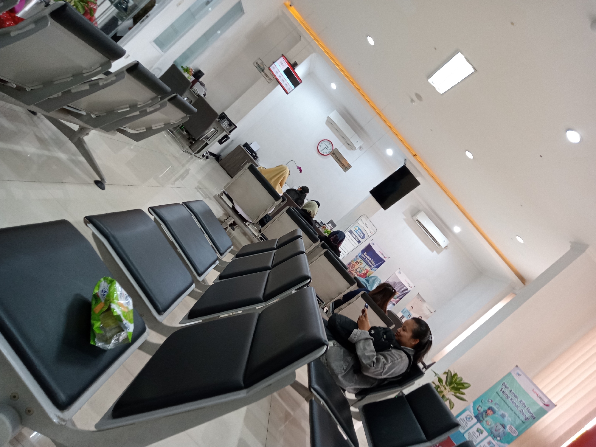 The image depicts a waiting area with rows of black, padded chairs. In the foreground, a green, crumpled snack wrapper rests on one of the seats. To the right, a woman in a grey jacket is seated, looking at her phone. Further back, other individuals are also seated and appear to be waiting. The room has white walls and a tiled floor, with overhead lighting. Several signs and a clock are visible on the wall, and a black television screen is mounted on the ceiling. The overall impression is that of a clean, modern waiting space, possibly in a public building or office. Without additional context or landmarks specific to Batu, Indonesia, it is impossible to definitively pinpoint the location within the city.