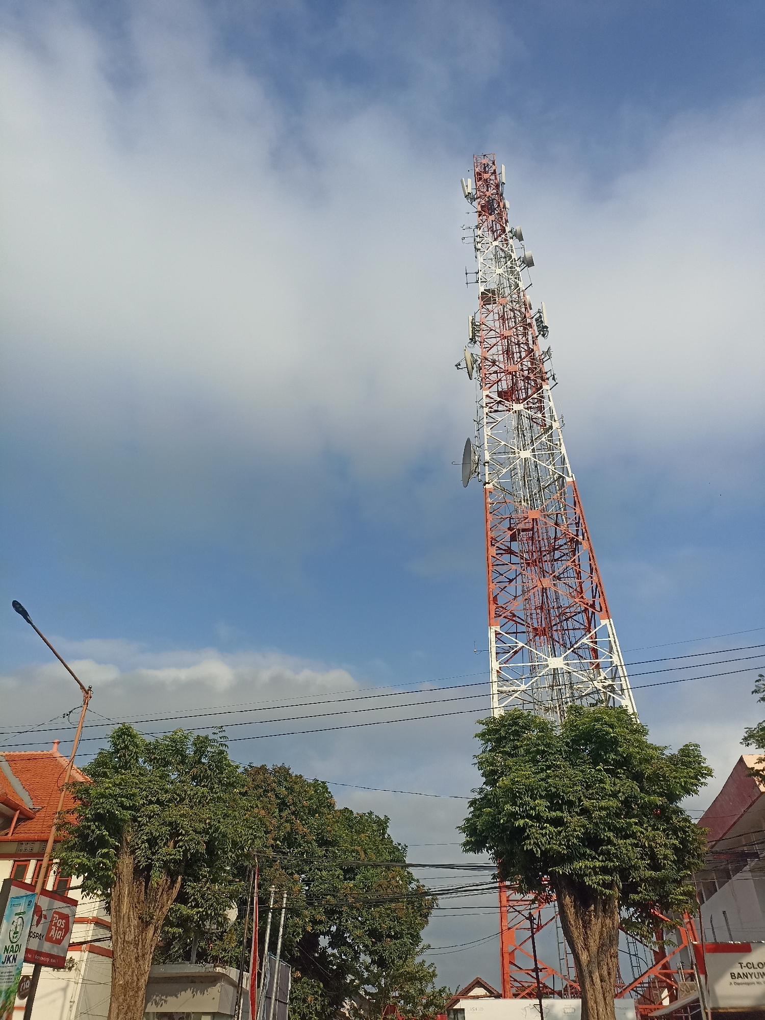 A tall, red and white telecommunications tower dominates the frame, angled slightly to the left and extending into a partly cloudy sky. The tower is a lattice structure with numerous antennas and satellite dishes attached. In the foreground, two large trees with dense green foliage stand at the base of the tower. To the left, a building with a red tiled roof and white walls is partially visible, adorned with signage including "NADI JKN" and "POS AJA!". A lamppost with a single light fixture extends from the left side of the image towards the center. Power lines are strung across the upper portion of the scene, connecting to poles. To the right of the tower, another building with a reddish-pink roof is partially visible. The ground is not clearly visible, but suggests an urban environment. The scene appears to be a street view in Kelurahan Kepatihan, Indonesia, likely during the daytime. There are no people visible in the image.