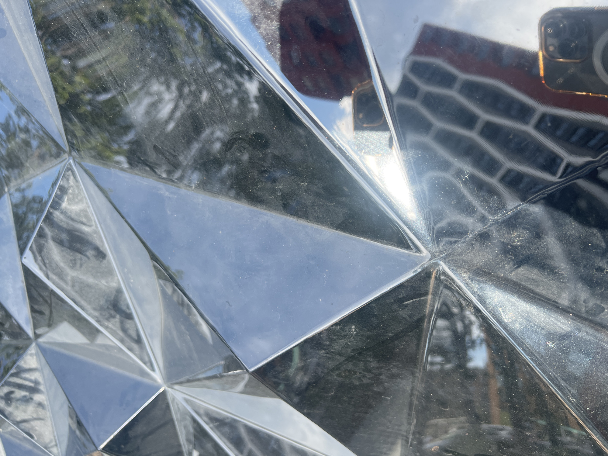 The media file captures a close-up view of a faceted, reflective surface, likely made of polished metal or glass. The surface is not smooth but composed of numerous geometric planes that catch and distort light, creating a mosaic-like pattern. These planes reflect the surrounding environment.
In the reflections, we can discern elements of an outdoor scene. Trees with green foliage are visible, suggesting it's daytime and likely spring or summer. There are also reflections of a building with a distinctive architectural pattern, possibly a facade with repeating hexagonal or grid-like elements. A bright sky is also reflected, indicating fair weather. A hand holding a smartphone is clearly reflected, with the phone's camera module visible. The phone has a gold-colored trim around the camera lenses. A dark-colored object, possibly a piece of clothing, is also reflected in another area of the faceted surface.
The overall impression is one of an urban or semi-urban setting where art or architecture features this highly reflective, geometric material. The lighting suggests it is daytime, possibly mid-morning or afternoon, given the bright highlights and shadows created by the sun. The specific location is Yekaterinburg, Russia. No text is visible in the media file.