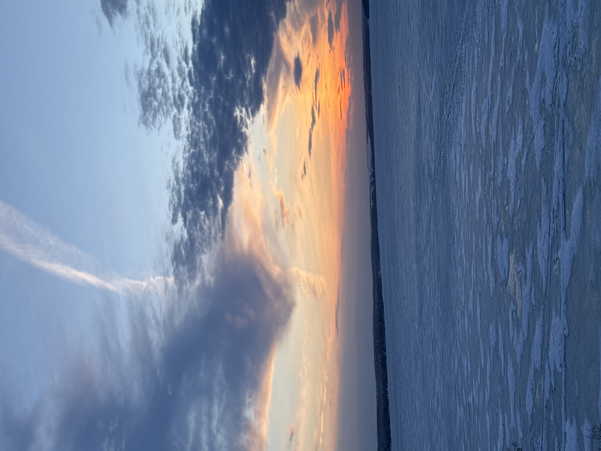 The image depicts a frozen body of water, possibly a lake or river, adjacent to a city. The sky displays a dramatic sunset with a mix of dark, textured clouds and orange-yellow hues. A contrail can be seen streaking across the sky. The frozen water surface appears textured with patterns of ice and snow. The location is assumed to be in Votkinsk, Russia, as requested. No people or specific actions are visible. The interaction is that of the environment elements with each other.