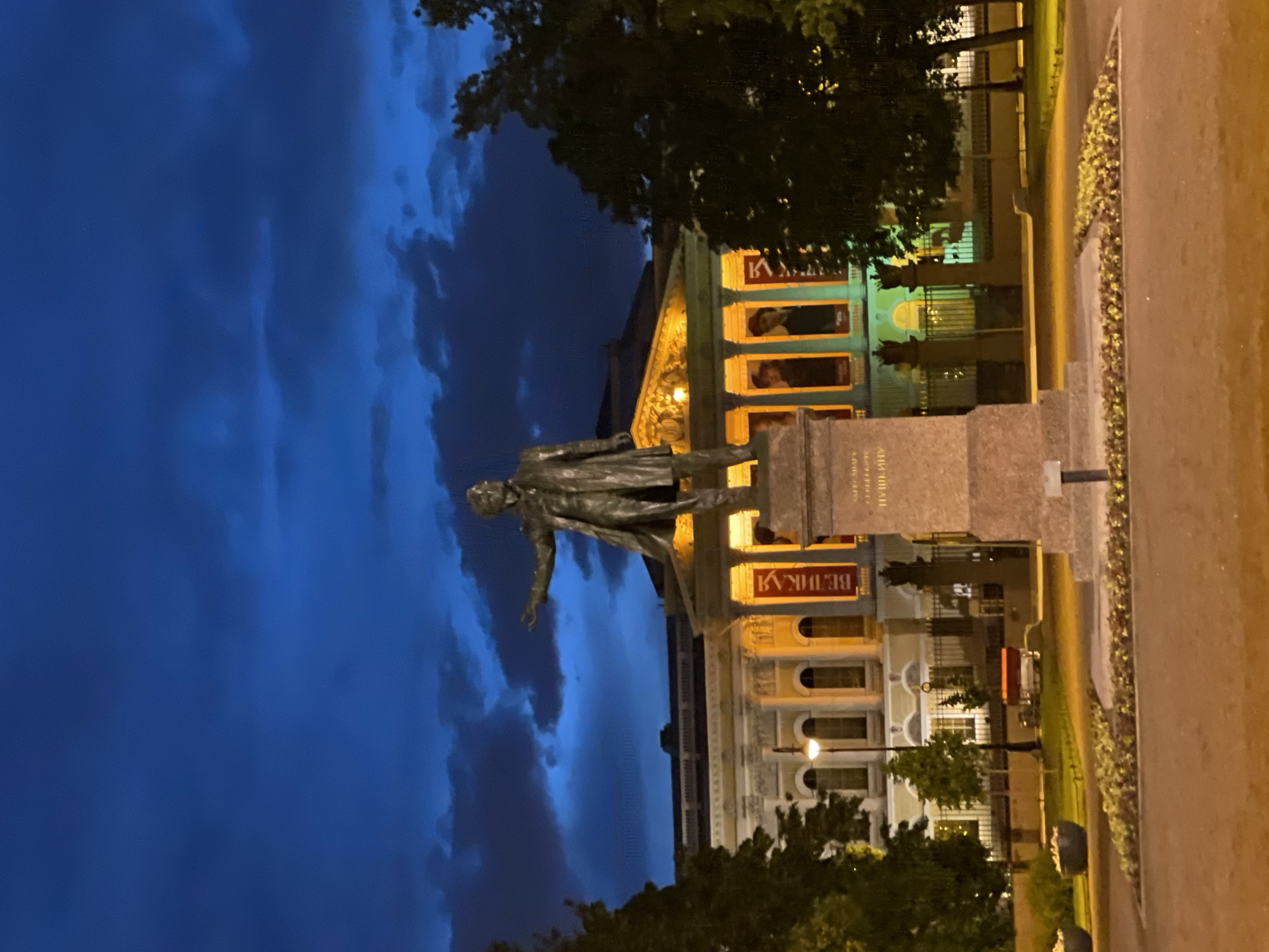 This nighttime image, rotated clockwise, depicts a statue of a historical figure in Saint Petersburg, Russia, set against the backdrop of a grand, illuminated building.
The central subject is a tall, dark statue of a man, likely made of bronze, standing on a rectangular stone pedestal. He is depicted wearing a long coat or cloak, with his left arm slightly extended. The pedestal bears Cyrillic inscriptions, which identify the figure as "А.С. ПУШКИН" (A.S. PUSHKIN) and state "РОЖДЕНИЯ 1799" (BORN 1799), confirming it is a monument to the renowned Russian poet Alexander Pushkin. Another visible word on the pedestal is "МИХАЙЛ" (MIKHAIL).
Behind the statue stands a large, ornate building with classical architectural elements, brightly lit with warm yellow light. Its facade features columns, arches, and intricate details in the pediment. Several large rectangular banners or illuminated windows on the building display imagery and text. One banner on the left partially shows "КАТРИН" (KATRIN), and another on the right clearly reads "ВЕЛИКАЯ" (VELIKAYA), suggesting an exhibition related to Catherine the Great. The building is likely the State Russian Museum, given the context of the Pushkin monument.
The sky above is a deep, dark blue, characteristic of twilight or night, with soft, visible clouds. Dark foliage from trees frames the scene on both sides and partially in front of the building. In the foreground, a paved path or road is visible on the right, alongside a narrow strip of greenery or flowerbed. The overall atmosphere is serene and majestic, highlighting the monument and architecture under artificial lighting.